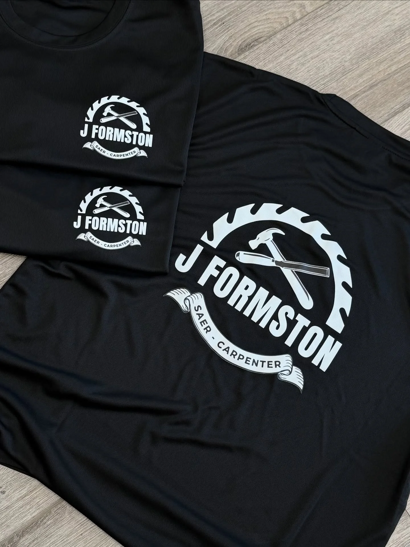 Black fabric with white logo featuring a saw blade, hammer, and tools, with the text 'J FORMSTON' and a ribbon beneath reading 'SAER - CARPENTER'.