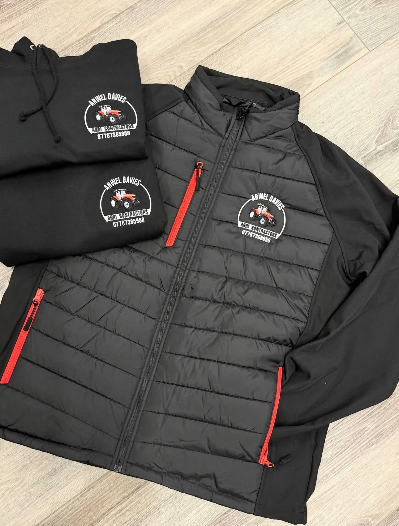 Black jacket with red zippers and a logo on the chest that reads "Arwel Davies Farm Contractors" with a tractor graphic, placed on a wooden floor.