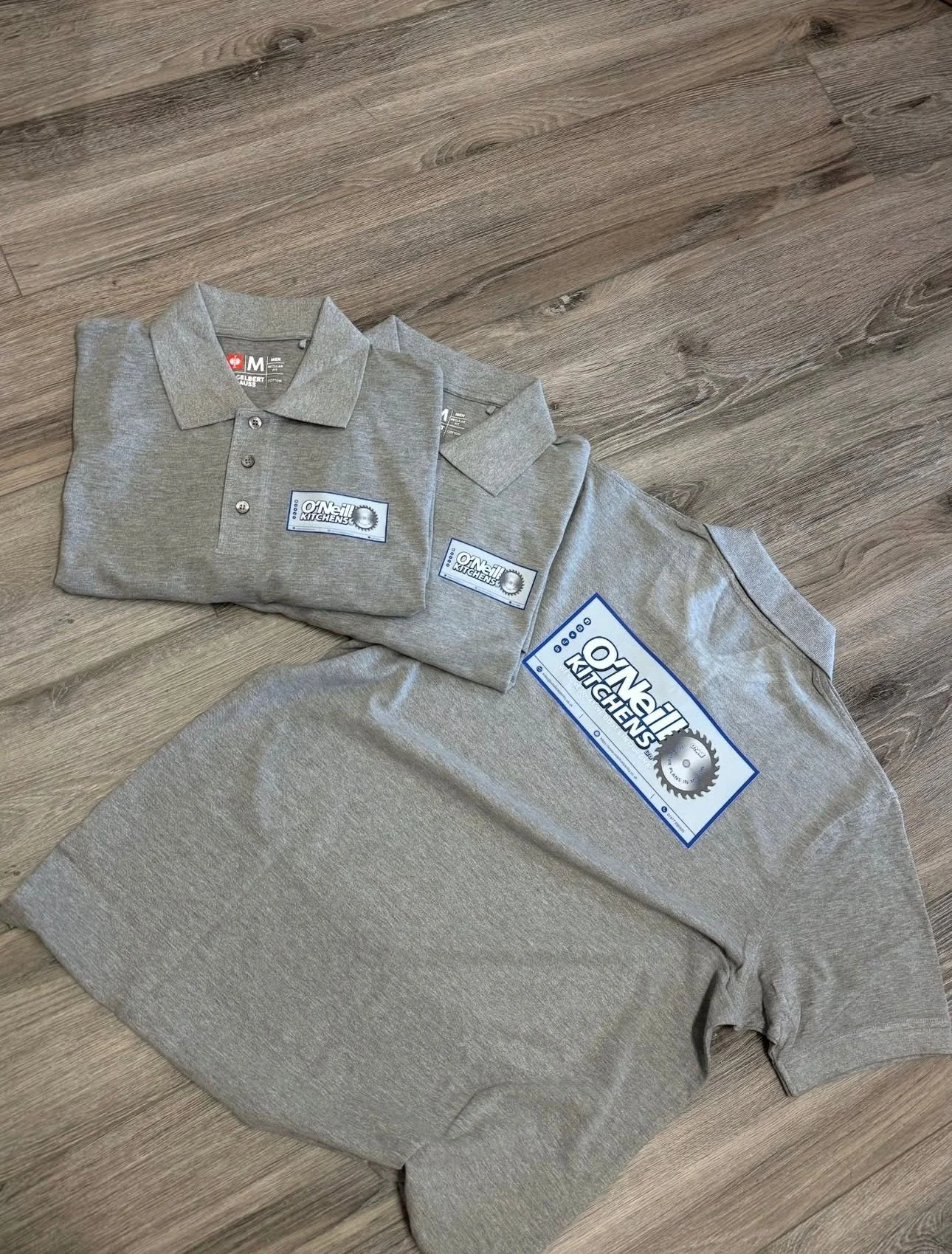 Gray polo shirts and a gray sweatshirt with 'O'Neil Kitchens' logos on the front and sleeve, lying on a wooden floor.