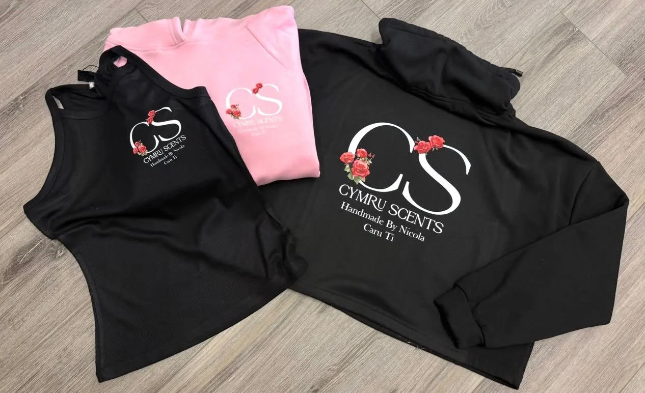 Three sweatshirts on a wooden floor, two black and one pink, with 'Cymru Scents' logo featuring red roses and text that reads 'Handmade By Nicola Caru Ti'.