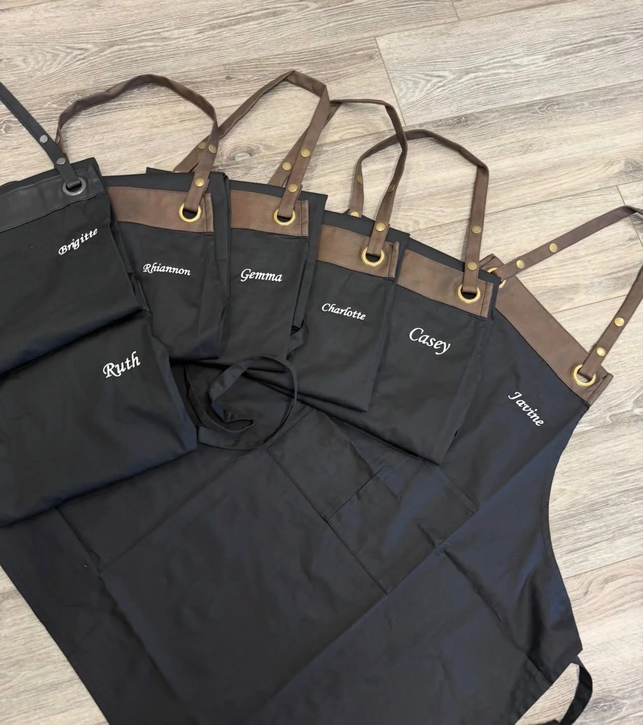 Personalized black apron and matching tote bags with brown straps and gold rings, each bag embroidered with a different name including Ruth, Rhiannon, Gemma, Charlotte, Casey, and Tairine, laid out on a wooden floor.