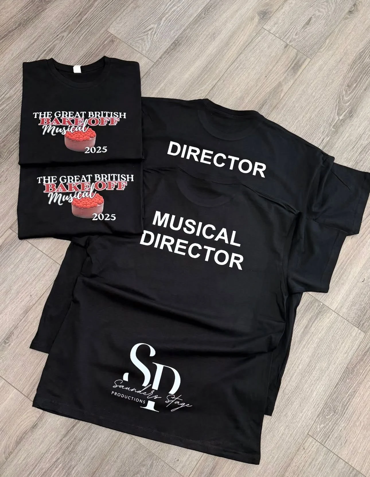 Black T-shirts with rehearsal and production crew names and titles. Some shirts feature the logo for 'The Great British Bake Off Musical 2025' with a cake image. Other shirts display roles such as 'Director' and 'Musical Director' in large white text