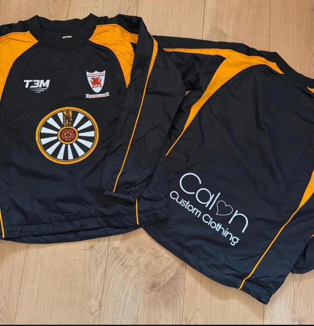 A black and yellow sports jacket with a logo of Johnstown Football Club, the letters T3M, and a circular emblem featuring a lion and a red rose. The back of the jacket has a white "Calm Custom Clothing" logo with a heart shape.
