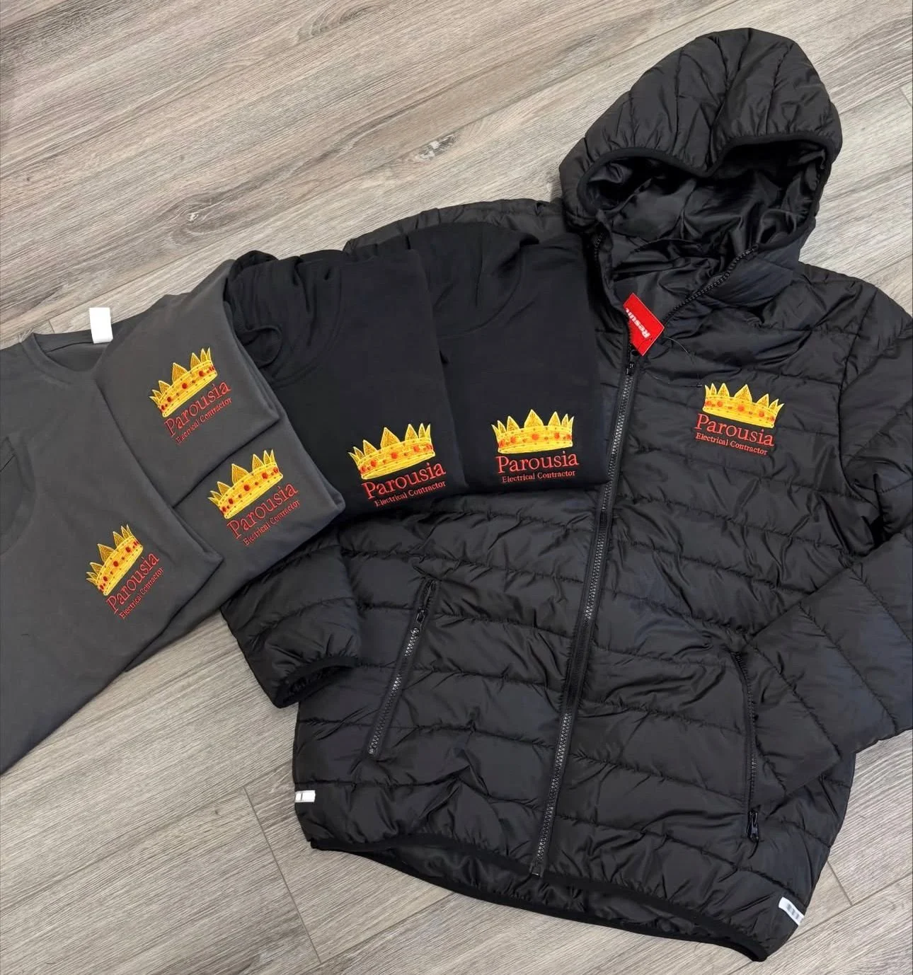 Black jacket and three gray and black t-shirts, all with a logo that reads 'Parousia Electrical Contractor' with a gold crown above the text, laid out on a wooden floor.