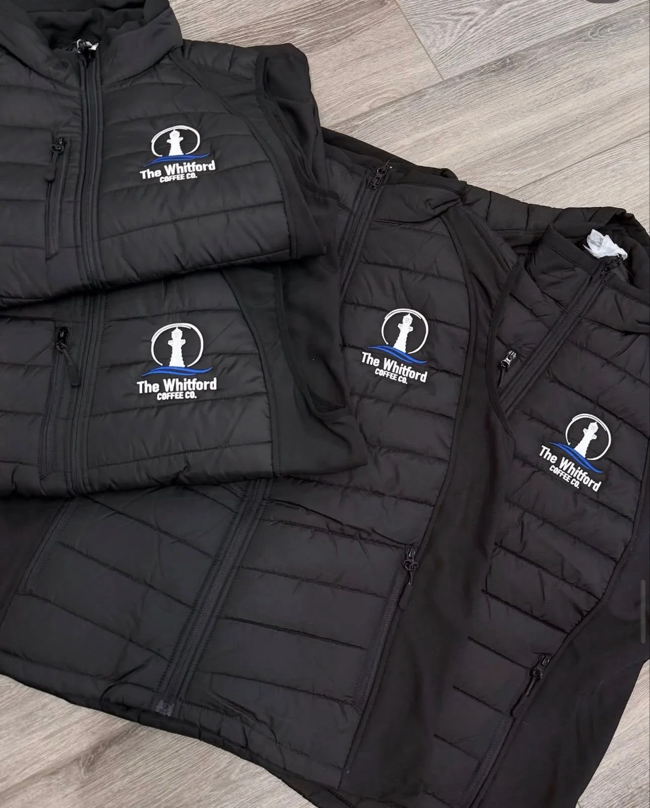 Black jackets with a logo for The Whitford Coffee Co. laid out on a wooden floor.