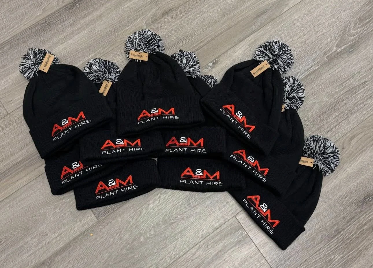 Eight black knit beanies with pom-poms, each embroidered with a red and white logo reading 'A&M Plant Hire,' are arranged on a wooden floor.