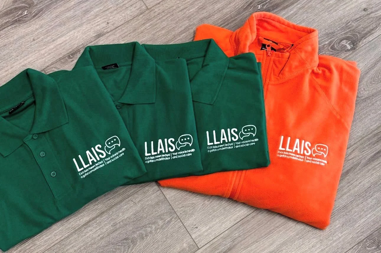 Three folded t-shirts, two green and one orange, with white text and a logo printed on them, placed on a wooden floor.