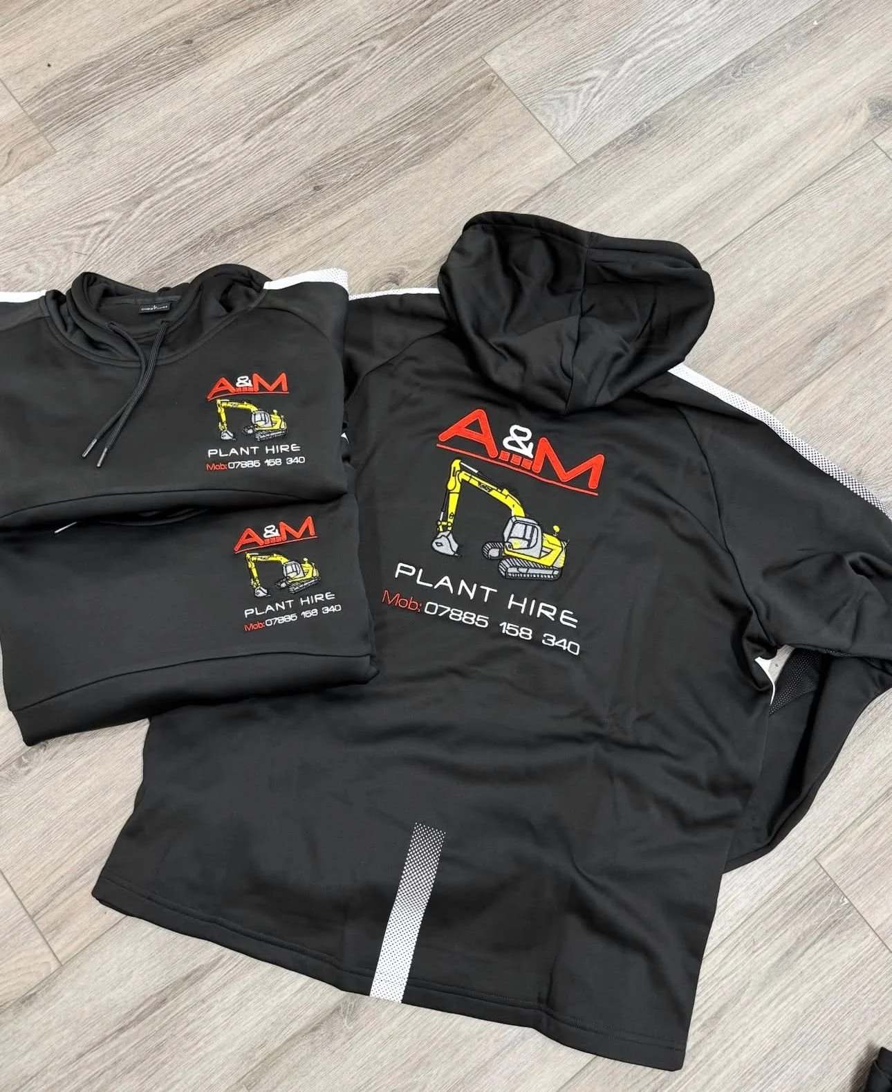 Black hooded sweatshirt, two black T-shirts, and a black jacket with a logo featuring a yellow excavator and the text 'A&M Plant Hire' and contact details, laid out on a light wood floor.