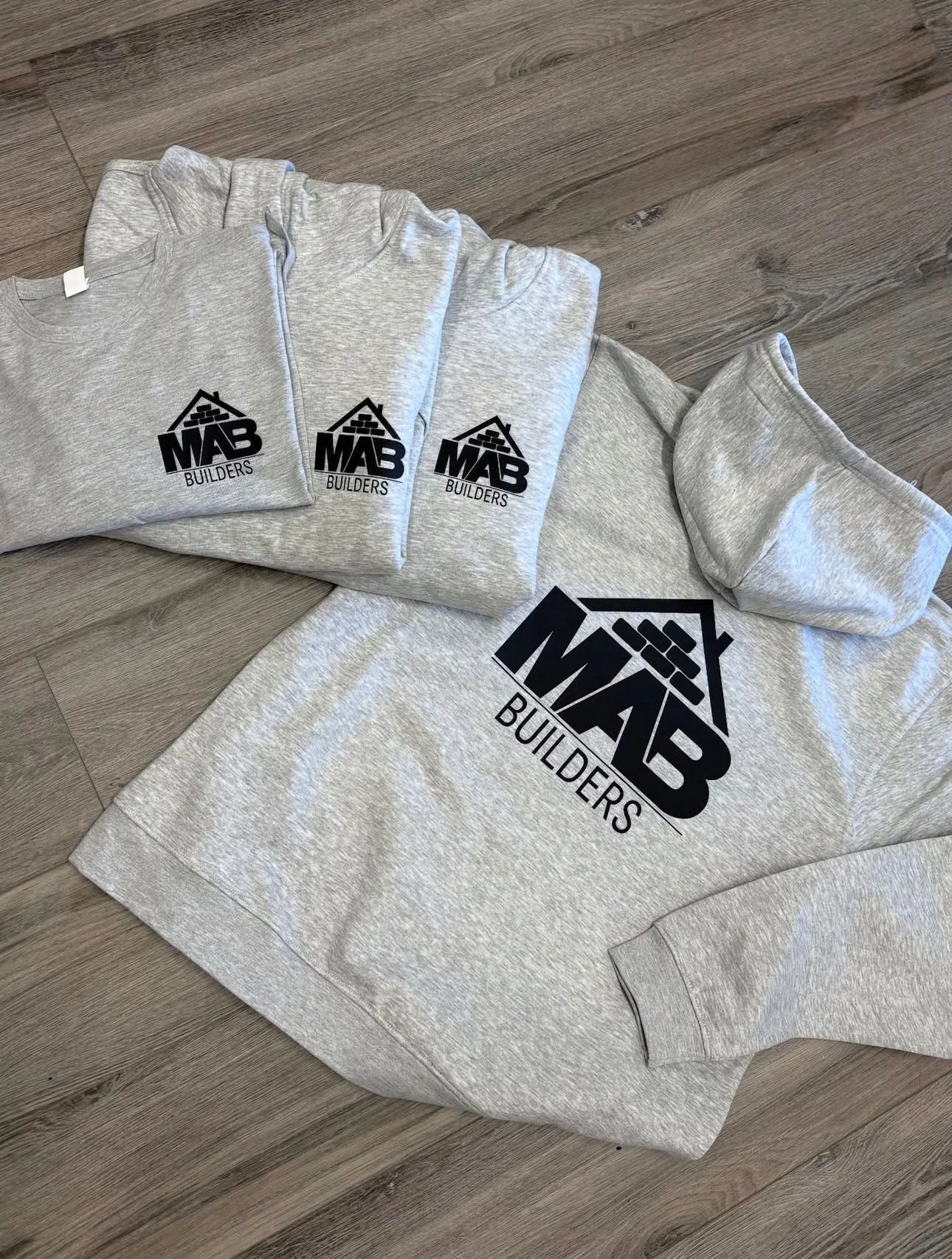 Gray sweatshirt, hoodie, and t-shirt with 'MAB BUILDERS' logo, laid out on a wooden floor.