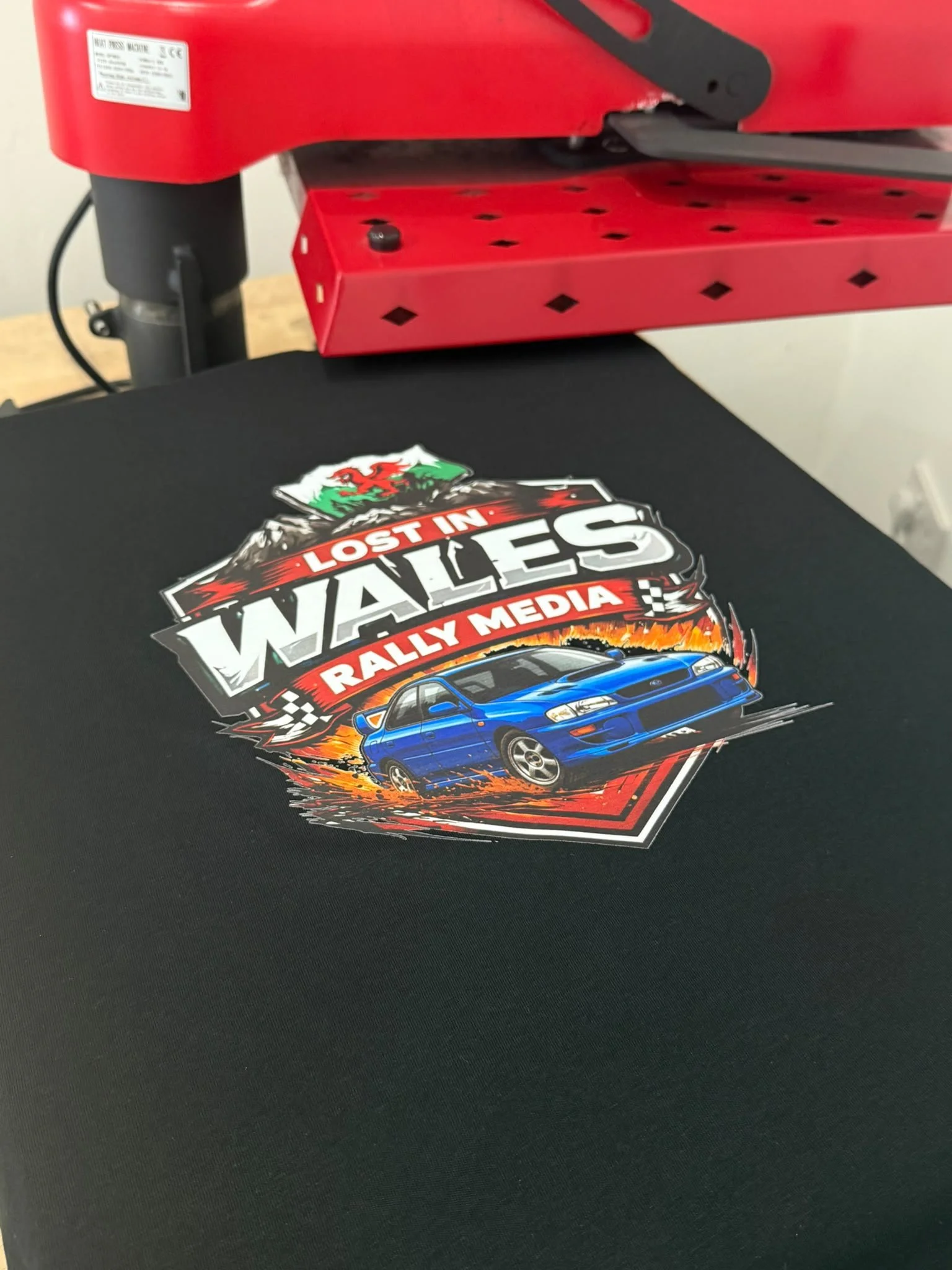 Close-up of a colorful sticker featuring a blue car, a Welsh flag, and the text "Lost in Wales Rally Media," on a black surface with a red mechanical device in the background.
