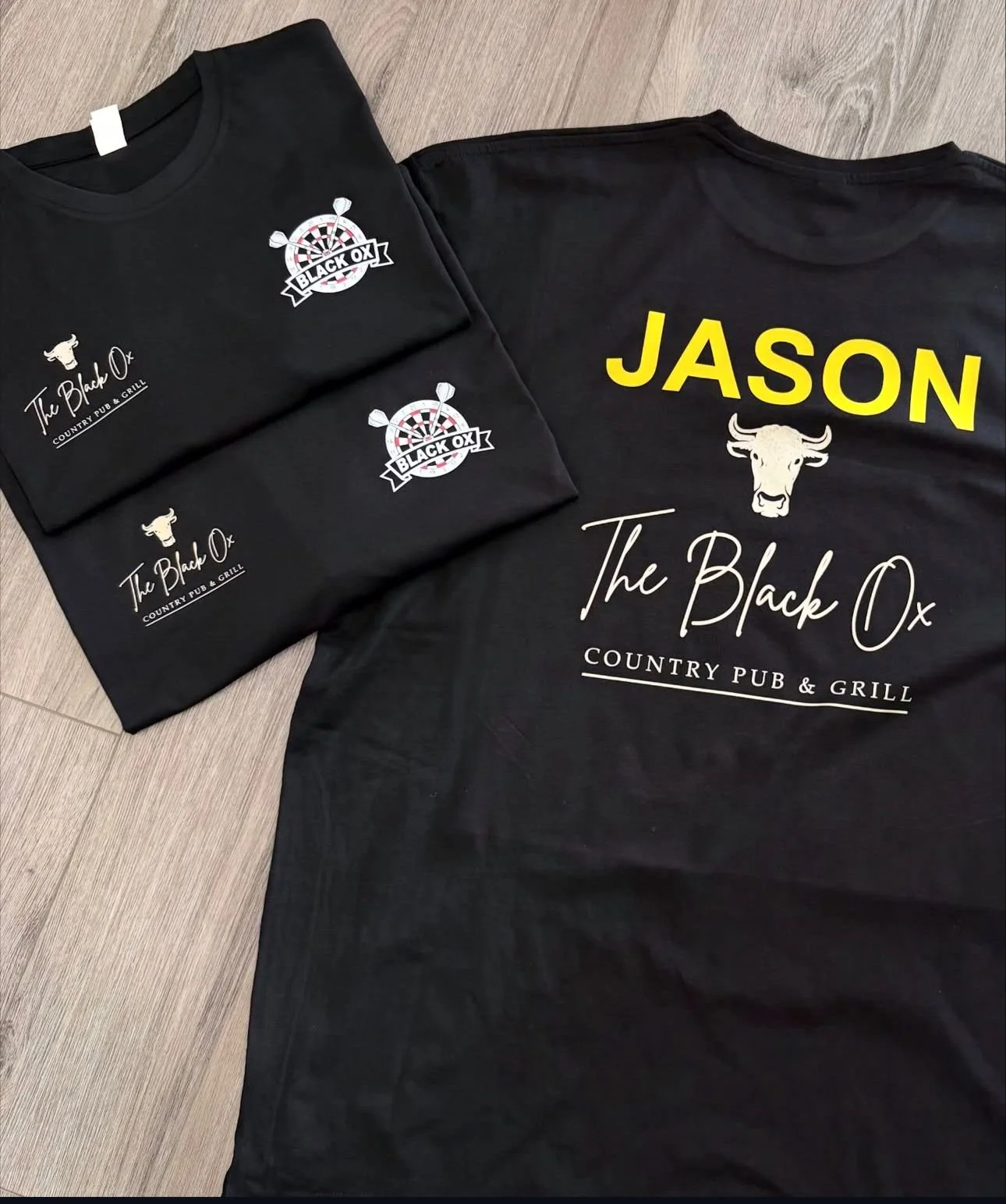 Group of black t-shirts and a black long sleeve shirt with logos and text for The Black Ox Country Pub & Grill, featuring a bull's head and a dartboard with the words "Black Ox".