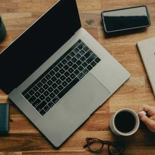 Paul Wogan Therapy. Top-down view of a workspace with a laptop, smartphone, notebook, coffee mug, and eyeglasses on a wooden desk.