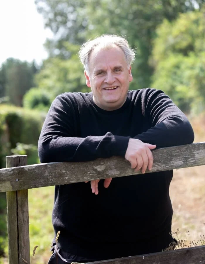 Paul Wogan smiling with gray hair wearing a black long-sleeve shirt leaning on a wooden fence outdoors with green trees and foliage in the background.