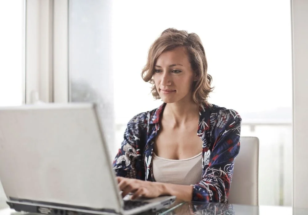 Paul Wogan Therapy. A woman with short, curly hair sits at a table using a laptop in a bright room with large windows.