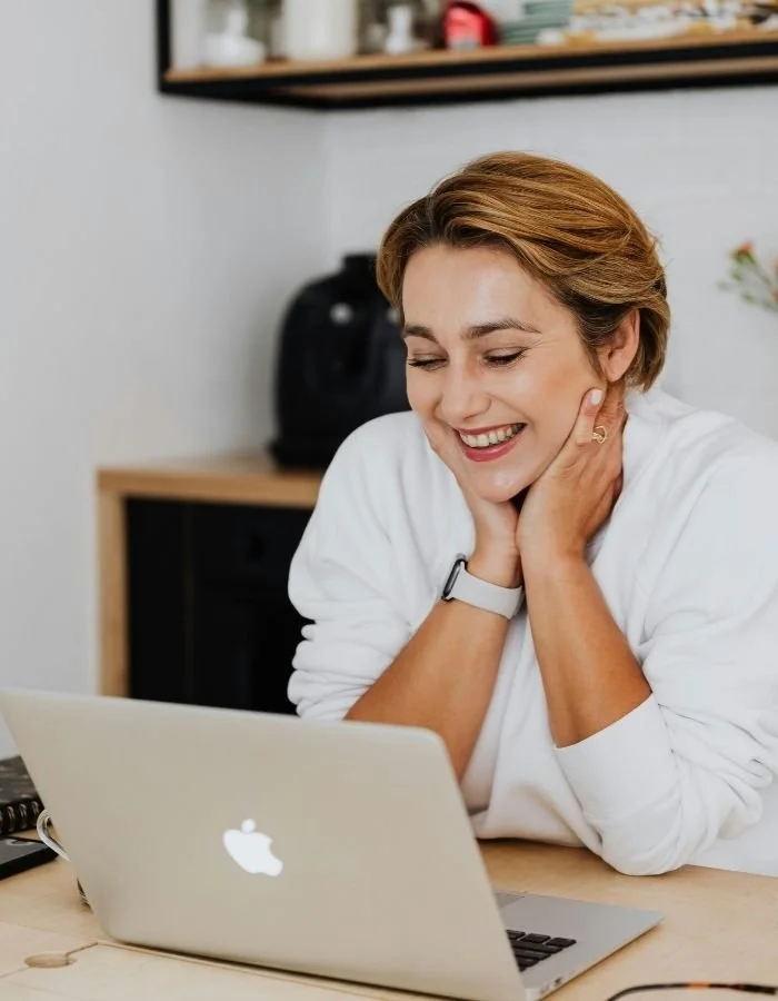 Paul Wogan Therapy. Woman smiling at her laptop in a modern office or home workspace.