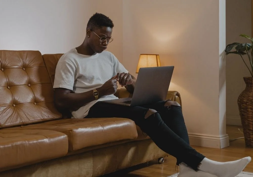 A person with short hair and glasses sits on a tan leather sofa in a living room, looking at a laptop. They are wearing a white shirt, ripped black jeans, white socks, and a watch. There is a small table lamp and a potted plant in the background.