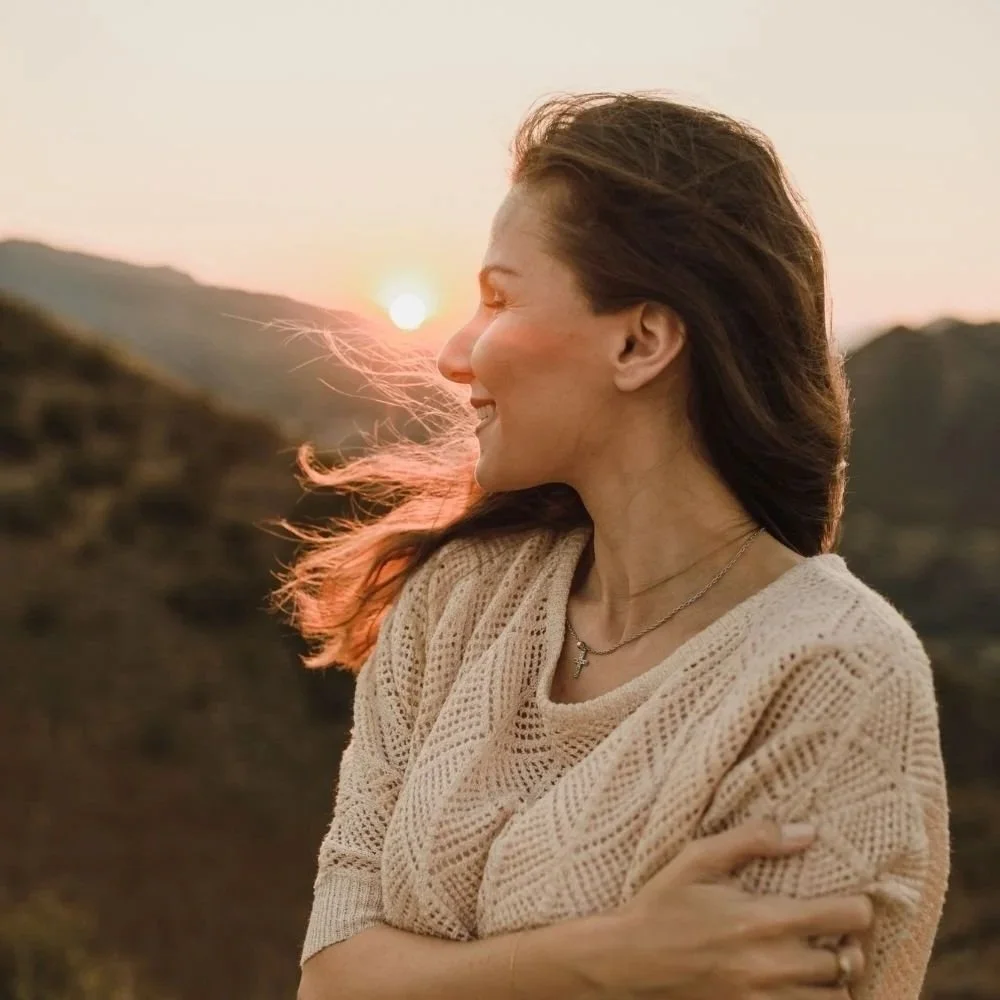 Paul Wogan Therapy. A woman with long hair smiling and standing outdoors during sunset, with hills in the background.