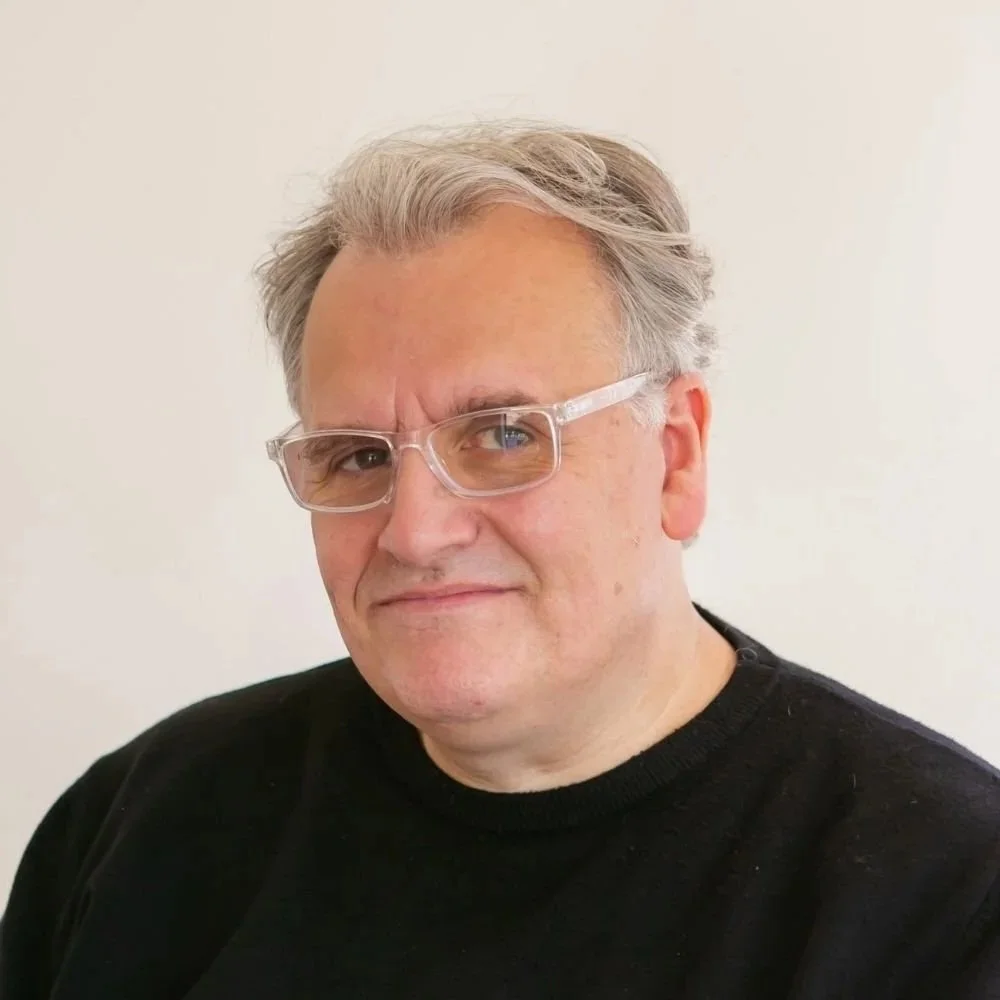 Paul Wogan with gray hair and glasses wearing a black shirt, standing against a plain white background.