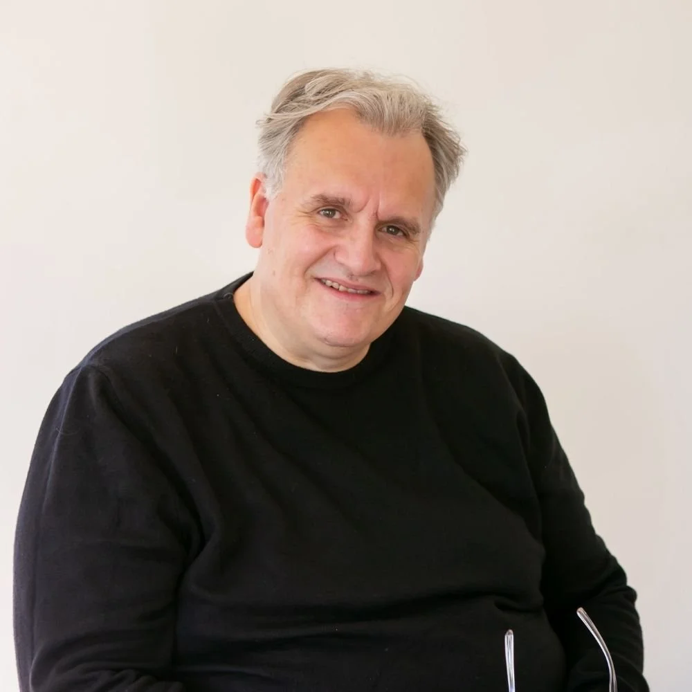 Paul Wogan with gray hair and a black sweater smiling at the camera against a plain light-colored background.