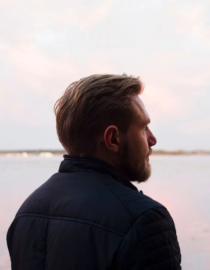 Paul Wogan Therapy. Profile of a man with styled hair and beard, wearing a dark jacket, overlooking a body of water during sunset or sunrise.