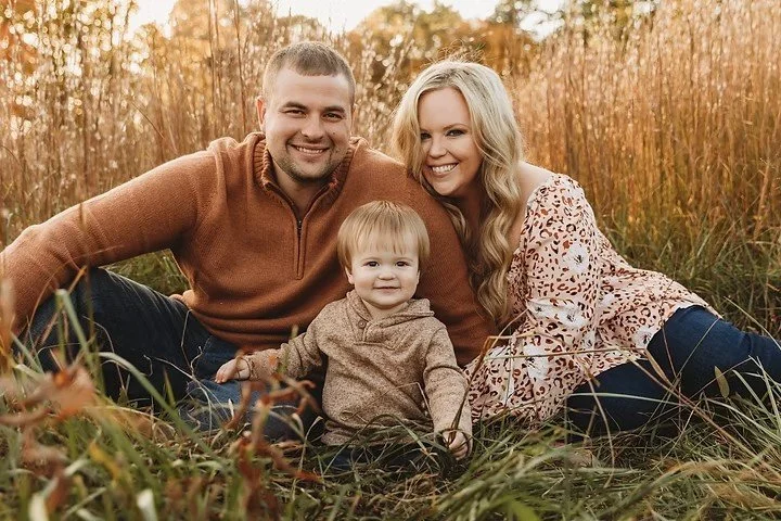 A happy family of three sitting on grass in a field during autumn, with trees in the background. The father, mother, and young child are smiling and posing for the camera.