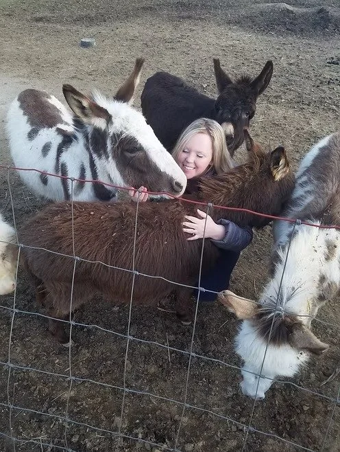 A young girl hugging a brown Donkey surrounded by several other miniature donkeys behind a wire fence on a dirt surface.