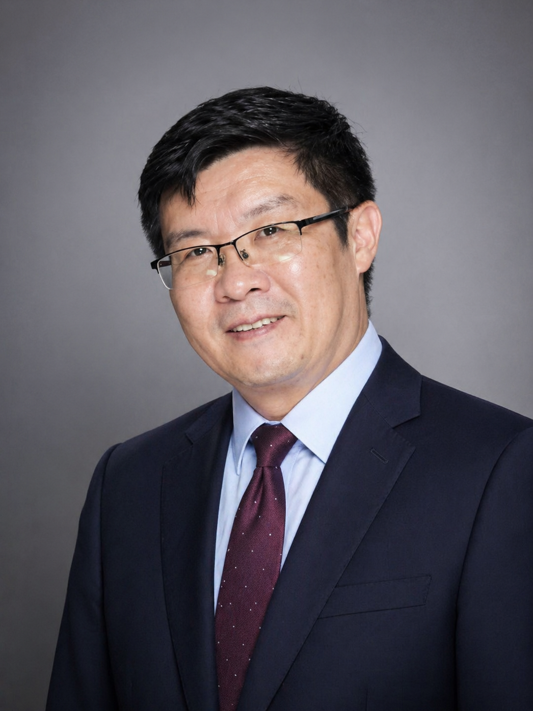 Headshot of an Asian man wearing glasses, a dark suit, light blue shirt, and a maroon tie, smiling against a gray background.