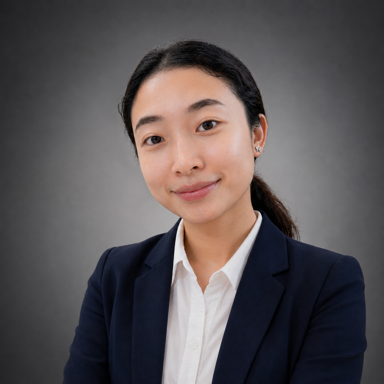A professional woman with dark hair tied back, wearing a navy blazer and white shirt, smiling at the camera against a gray background.