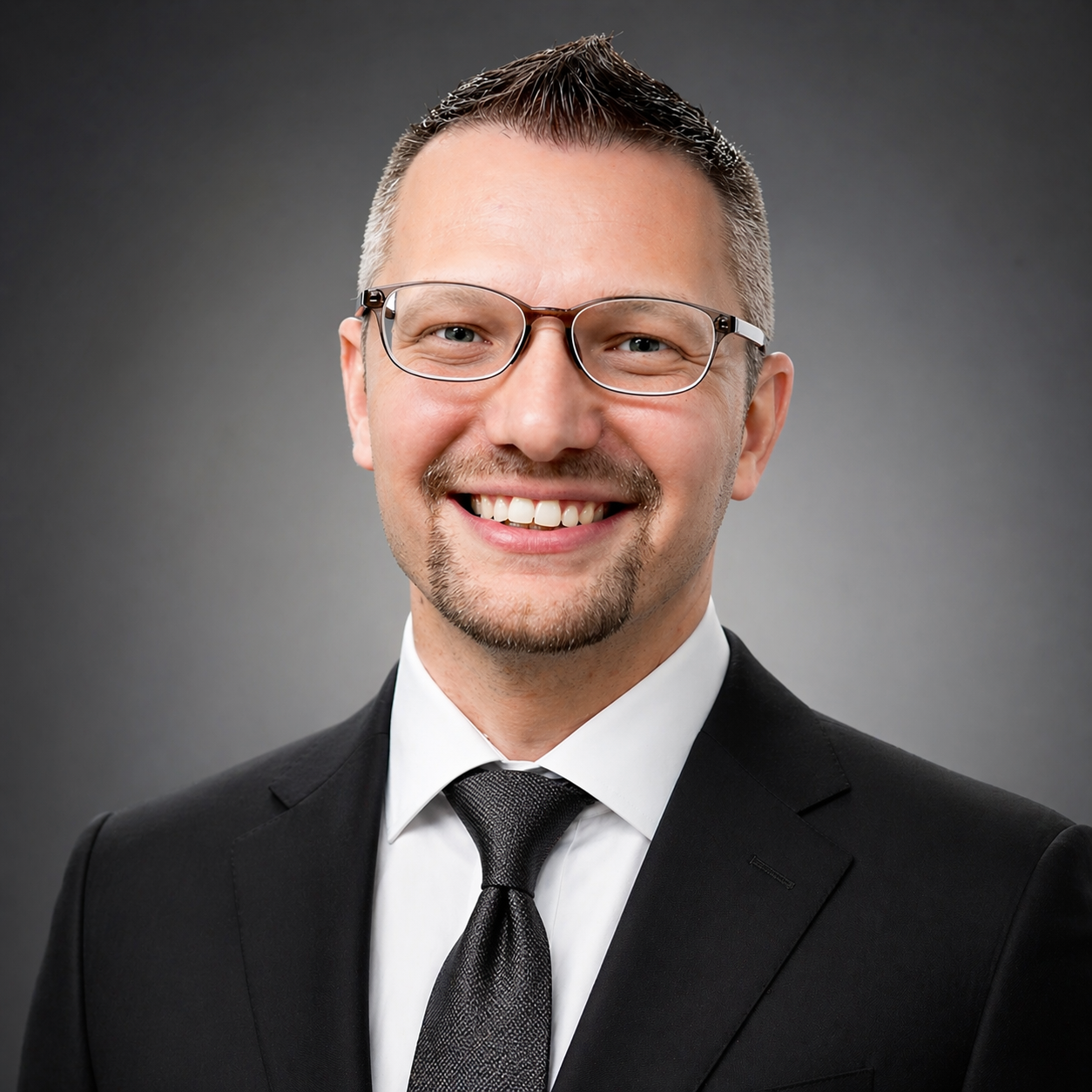 A smiling man wearing glasses, a black suit, white shirt, and black tie against a gray background.