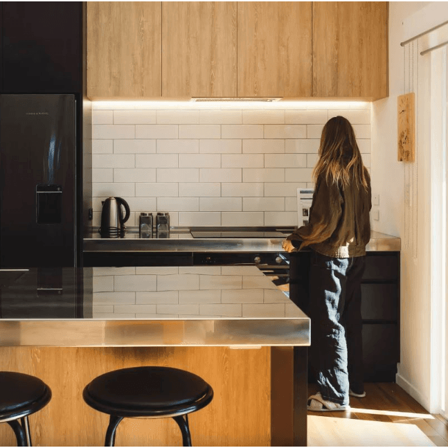 Adams Building architectural home in Fitzroy, New Plymouth with a woman with long hair wearing a brown jacket and jeans standing in a modern kitchen.