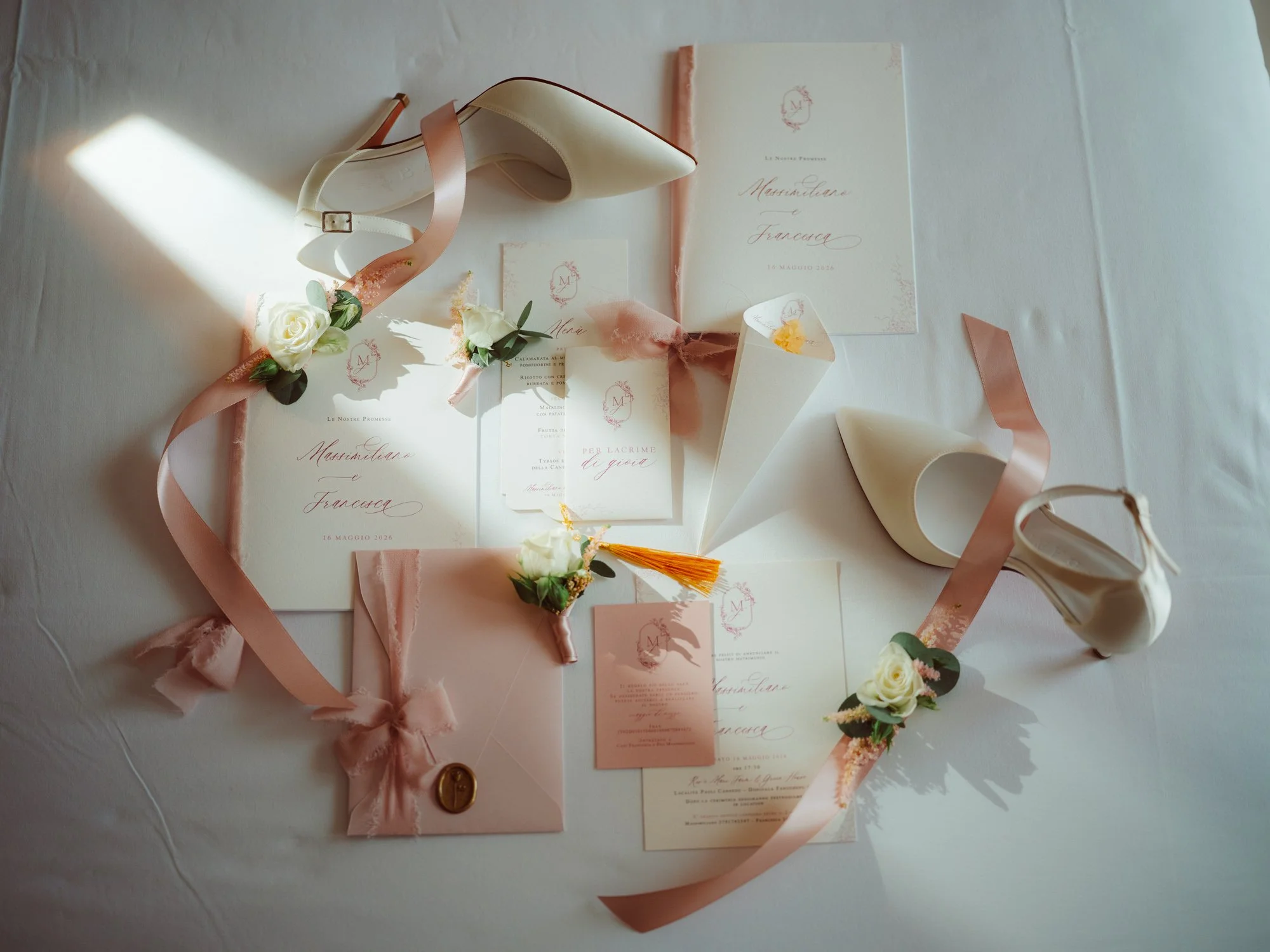 Flatlay con partecipazioni di nozze eleganti, scarpe da sposa color avorio, nastri in seta rosa e bottoniere