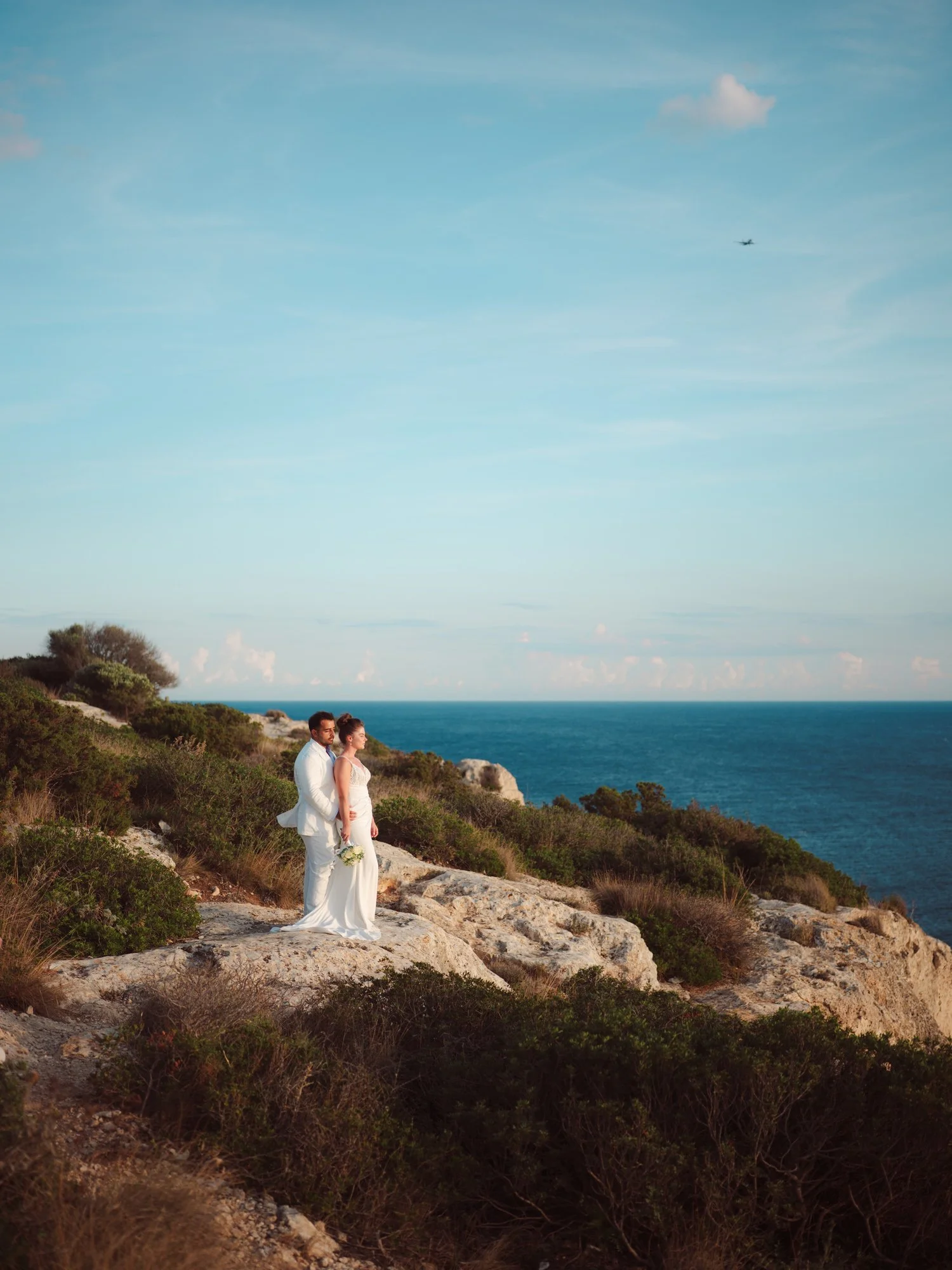 Una coppia di sposi in abiti eleganti si trova su una scogliera con vista sul mare, con cielo azzurro e nuvole sparse.