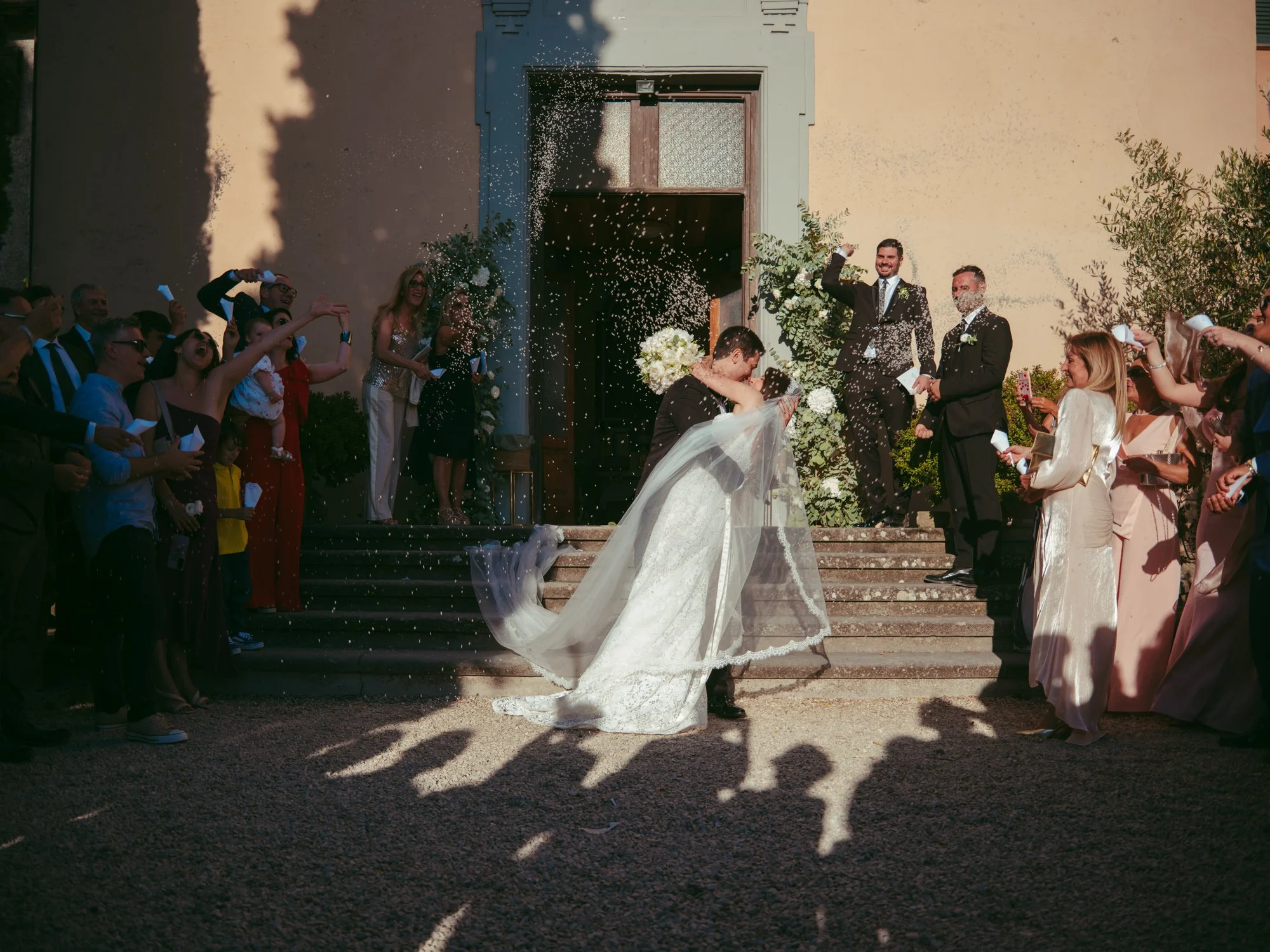 Gli sposi si baciano romanticamente sulla scalinata all'uscita dalla chiesa mentre gli invitati festeggiano lanciando il riso