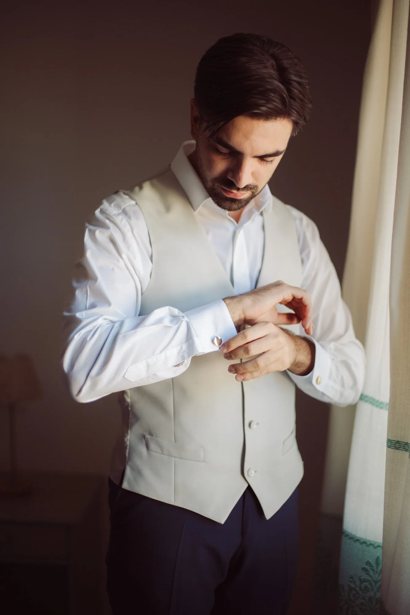 Primo piano dello sposo concentrato mentre allaccia i gemelli alla camicia bianca con gilet chiaro.