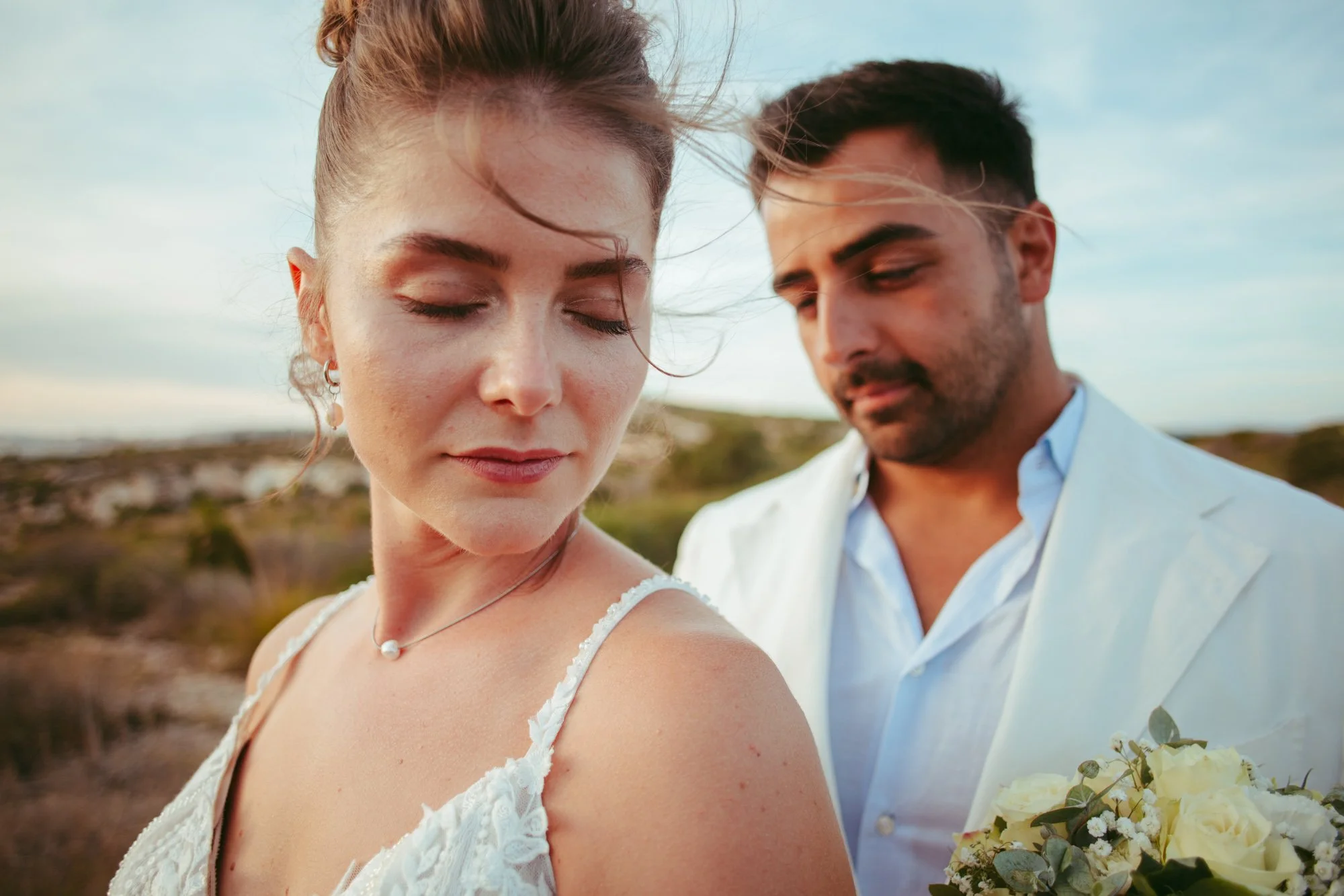 Primo piano delicato della sposa a occhi chiusi con il vento tra i capelli e lo sposo sfocato dietro di lei