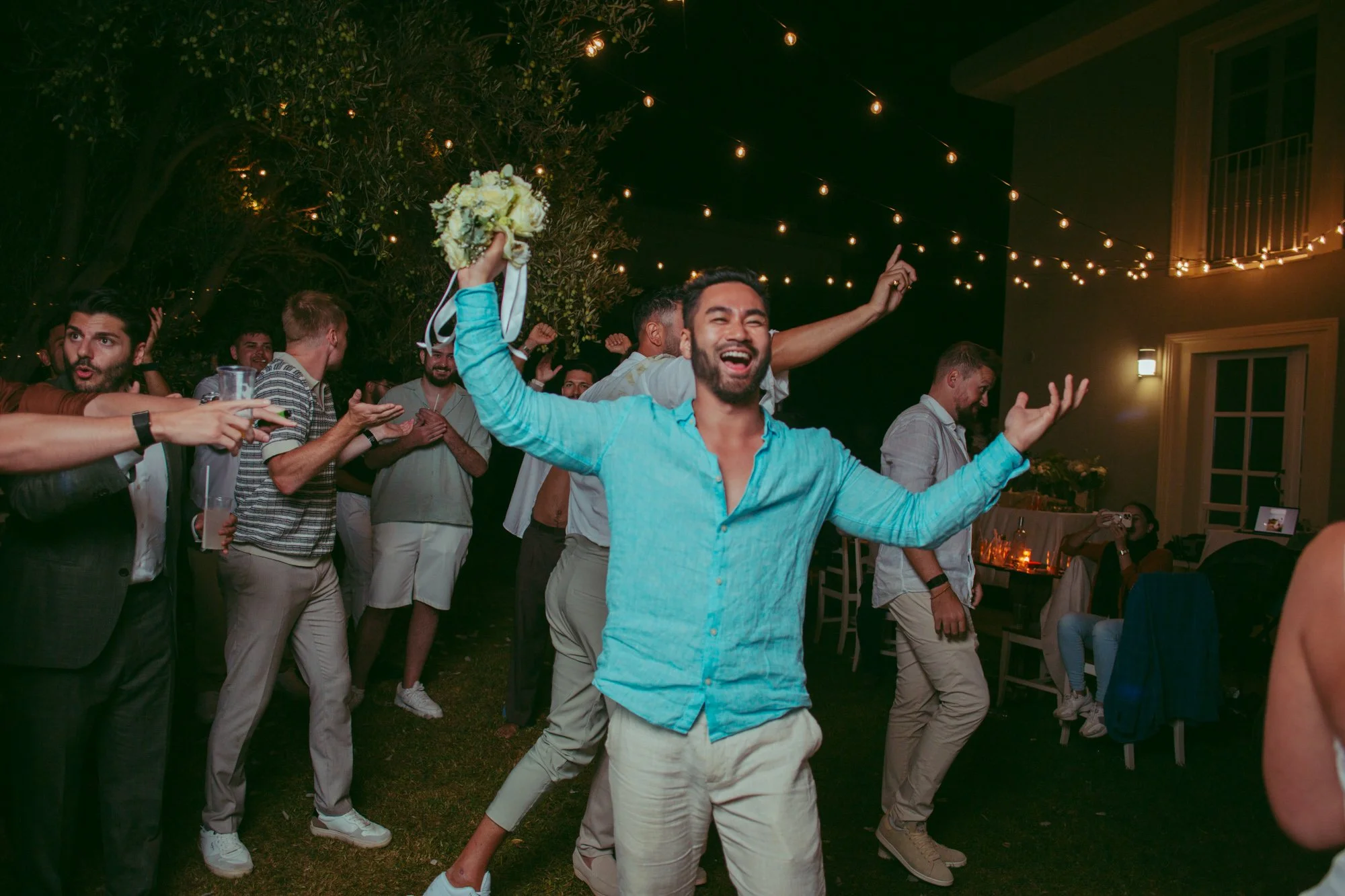 Un invitato sorridente in camicia azzurra alza il bouquet al cielo festeggiando la vittoria tra gli amici