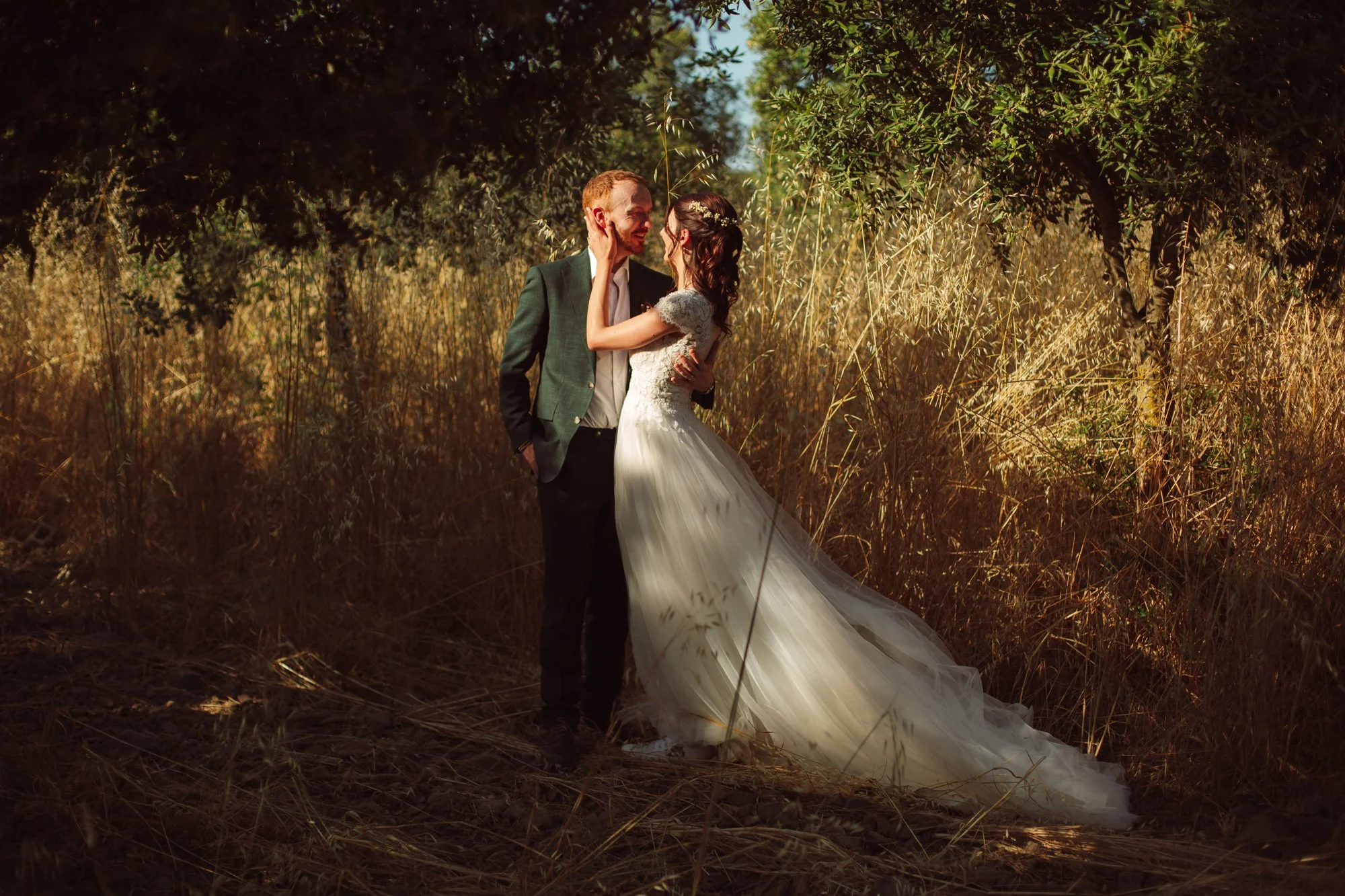 Una coppia di sposi in abiti da matrimonio si abbracciano e si sorridono in un bosco soleggiato con erba secca. La donna indossa un abito lungo e una coroncina di fiori, l'uomo indossa un soprabito verde scuro.