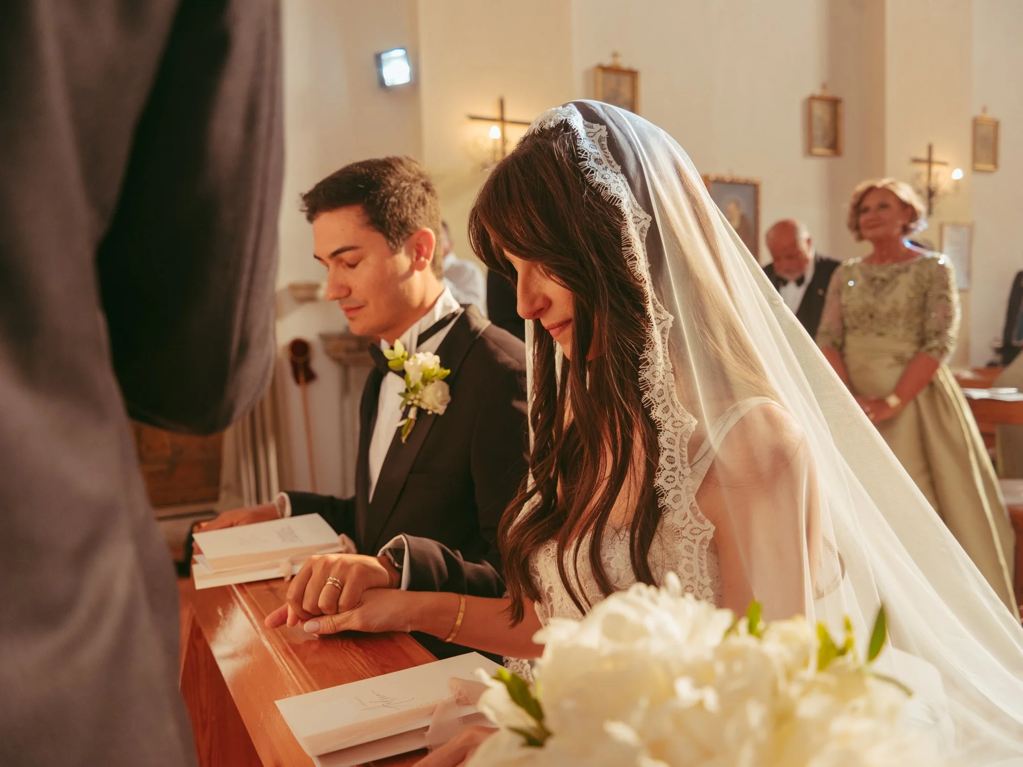 Gli sposi si tengono la mano sorridendo mentre sono posizionati all'inginocchiatoio durante la messa di nozze