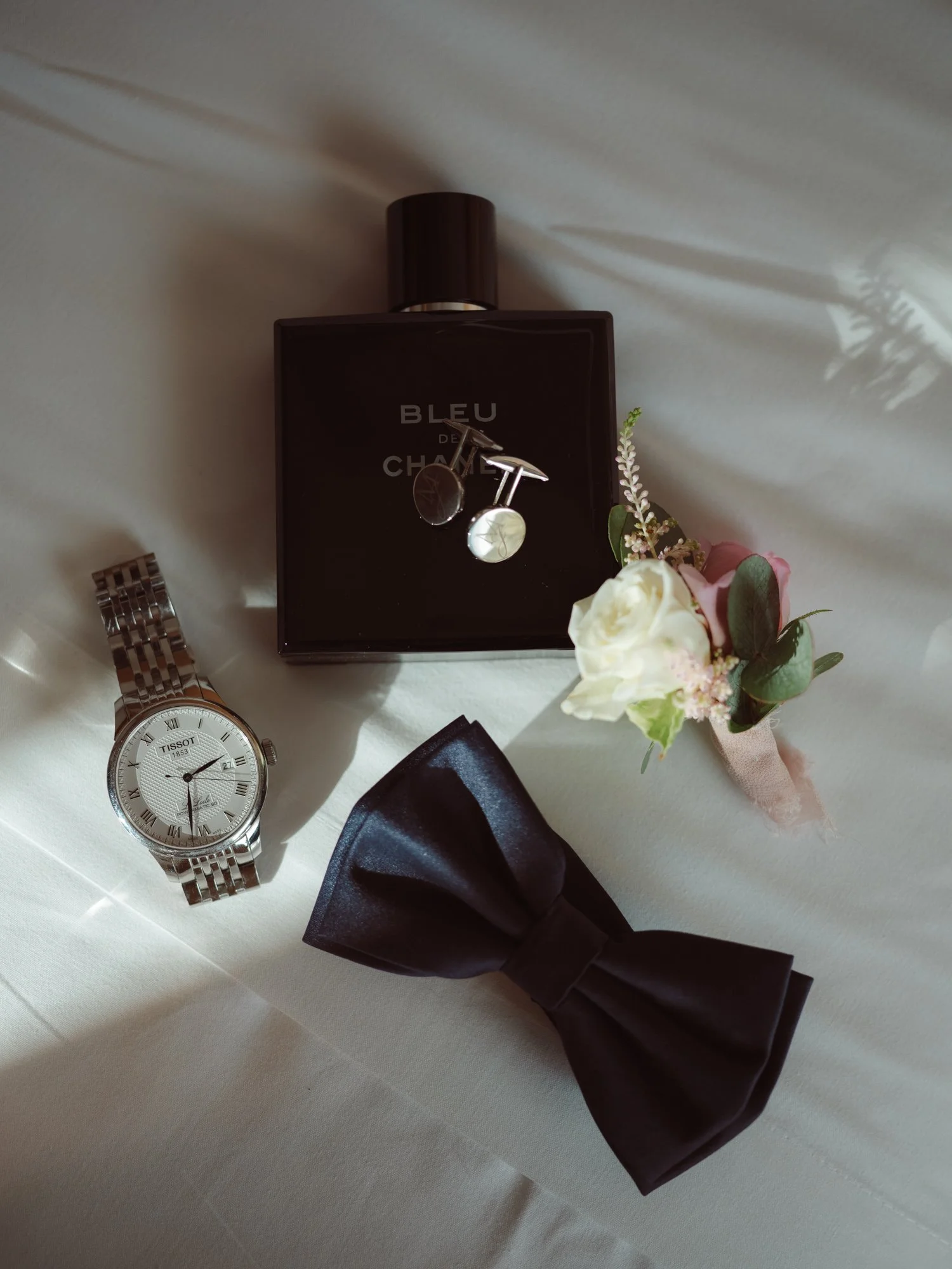 Flatlay con accessori dello sposo: profumo Bleu de Chanel, orologio Tissot, papillon scuro, gemelli e bottoniera con rosa