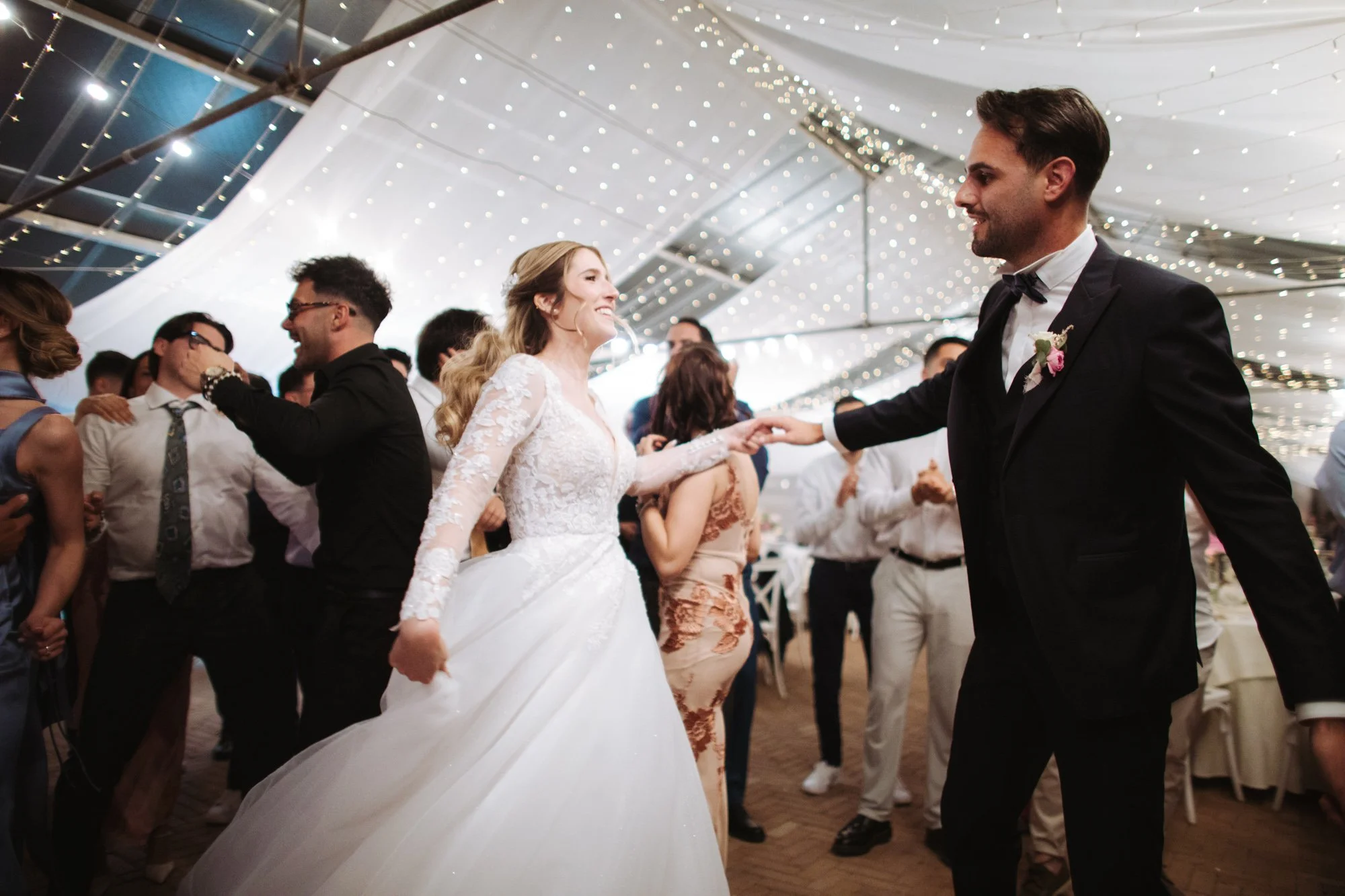 La sposa con abito bianco in pizzo gira tenendo la mano dello sposo mentre tutti intorno ballano
