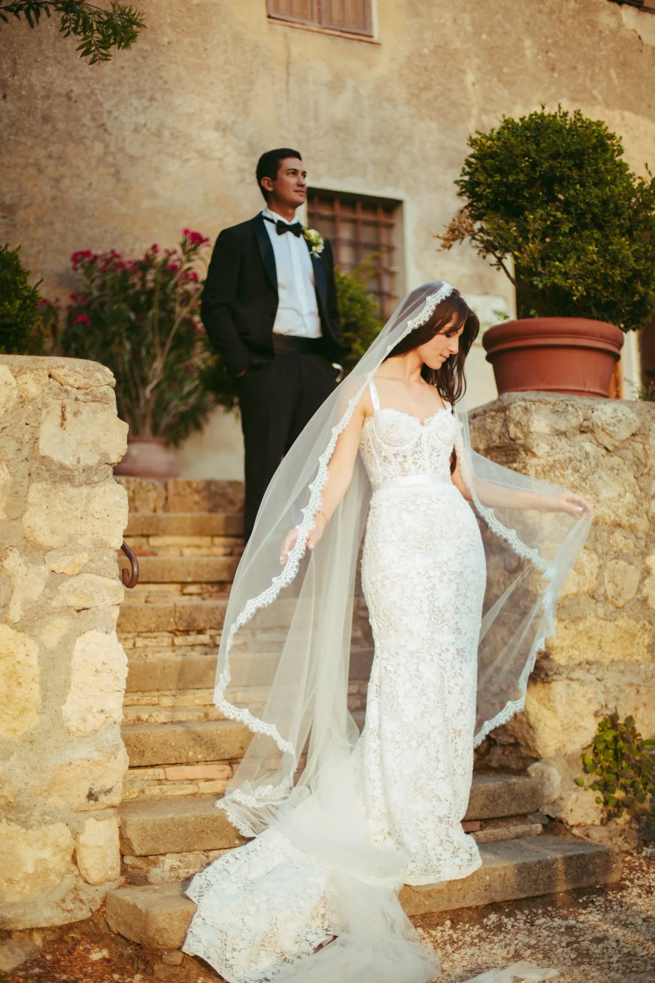 Ritratto della sposa in abito a sirena di pizzo in primo piano su una scalinata in pietra, con lo sposo in smoking sfocato sullo sfondo