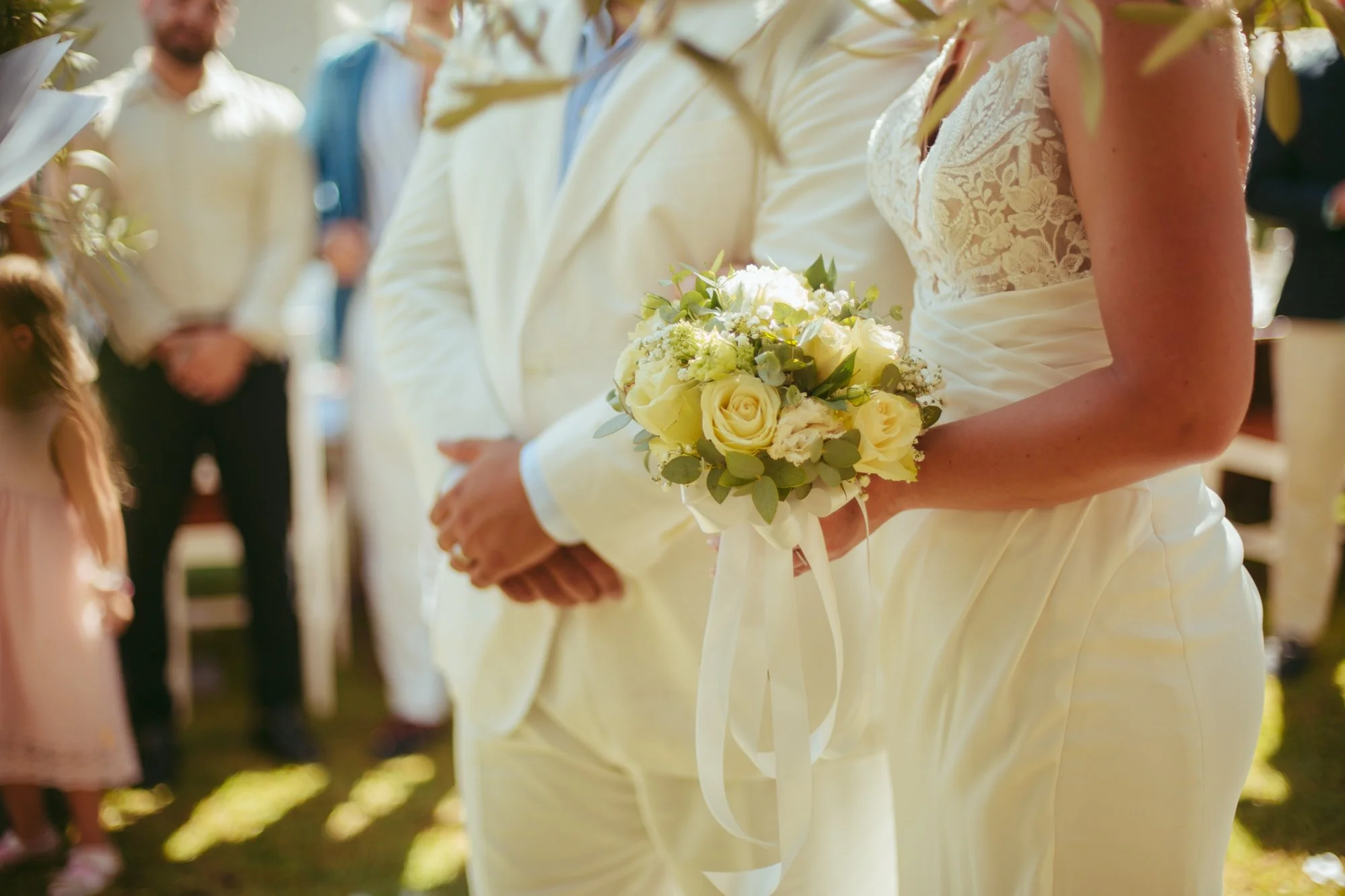 Dettaglio in primo piano delle mani della sposa che tiene un bouquet di rose bianche con nastro di seta accanto allo sposo