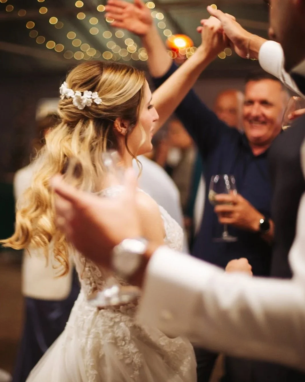 Coppia che danza in un matrimonio, con la sposa che indossa un abito bianco con ricami e una acconciatura con fiori, mentre un uomo sorride e tiene un bicchiere di vino.