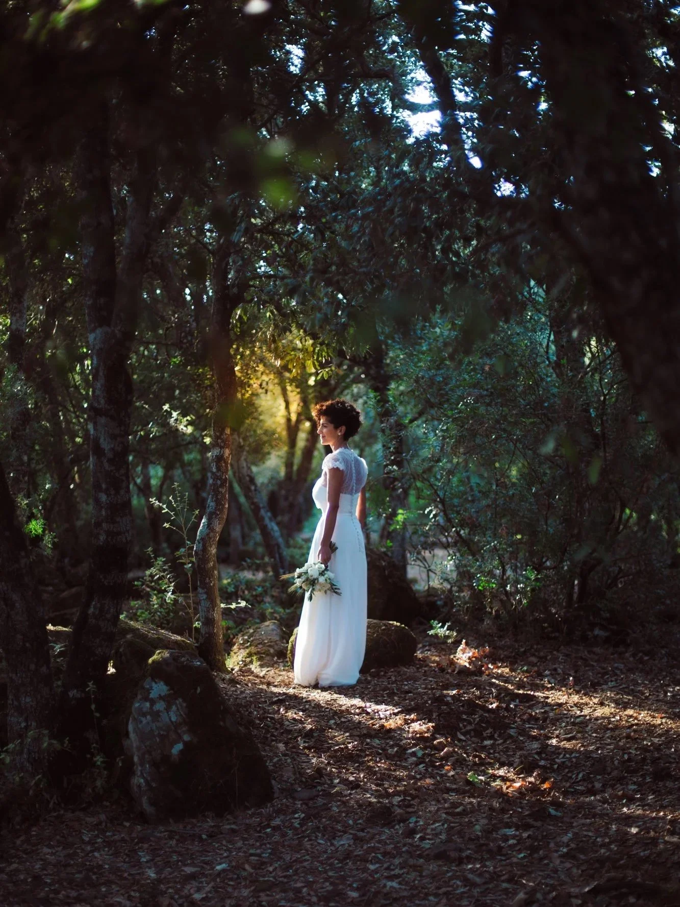 Una donna in un abito bianco da sposa che tiene un bouquet di fiori, si trova in un bosco con alberi e luce solare che filtra attraverso le foglie.