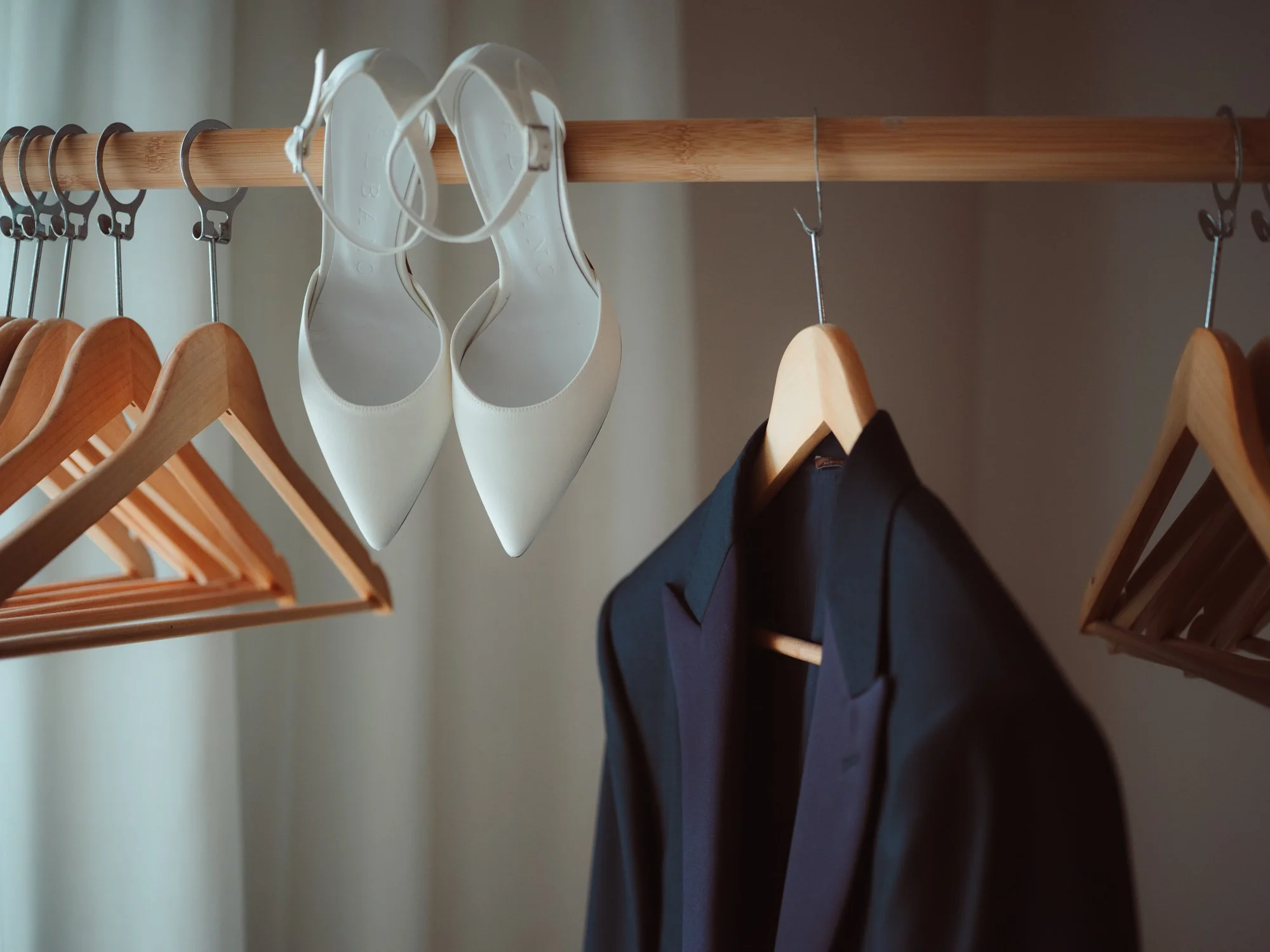 Scarpe da sposa con tacco bianche e giacca scura da sposo appese a uno stender in legno durante i preparativi