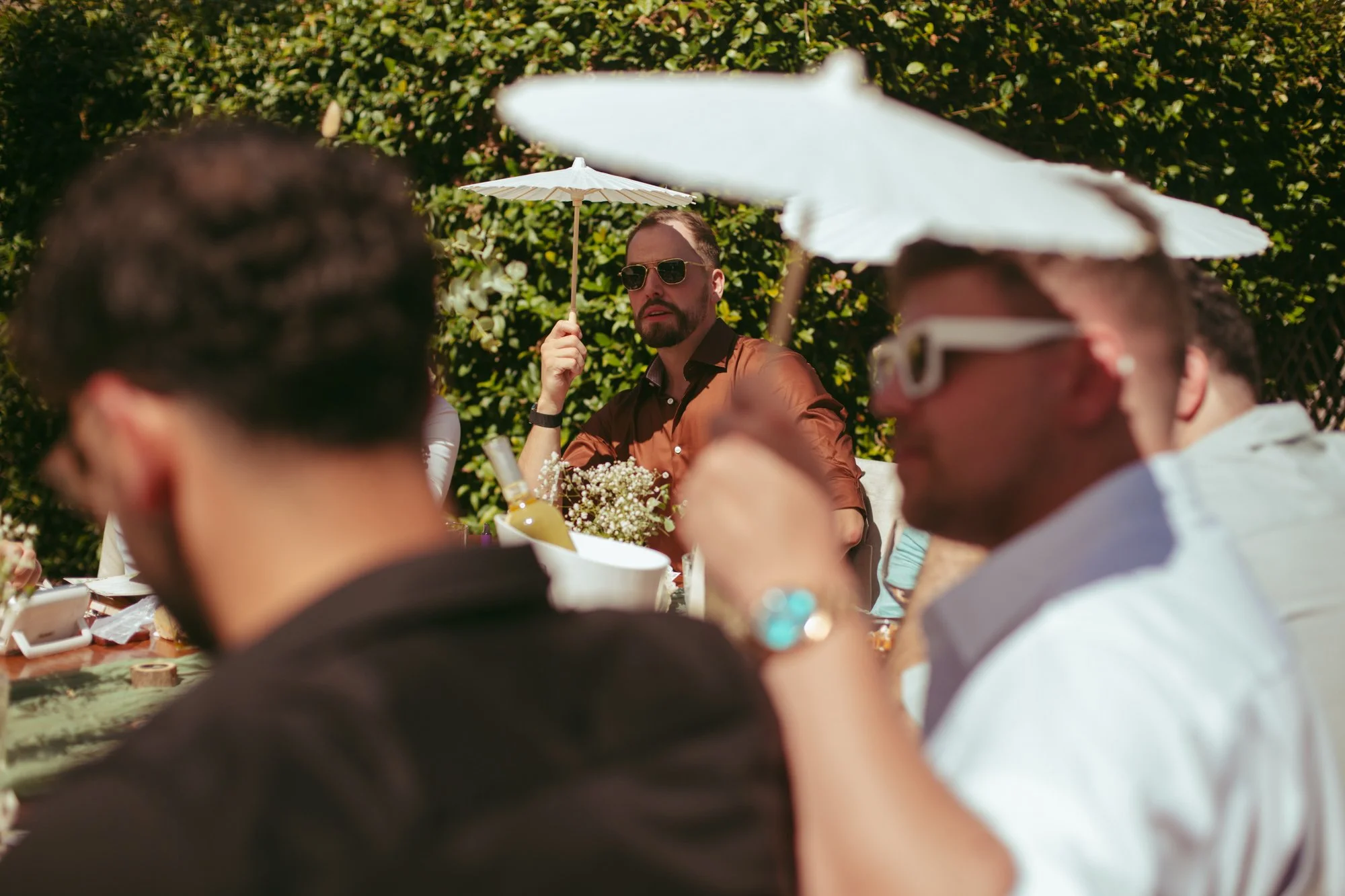 Un invitato con occhiali da sole tiene un piccolissimo ombrellino parasole bianco per ripararsi dal sole al tavolo