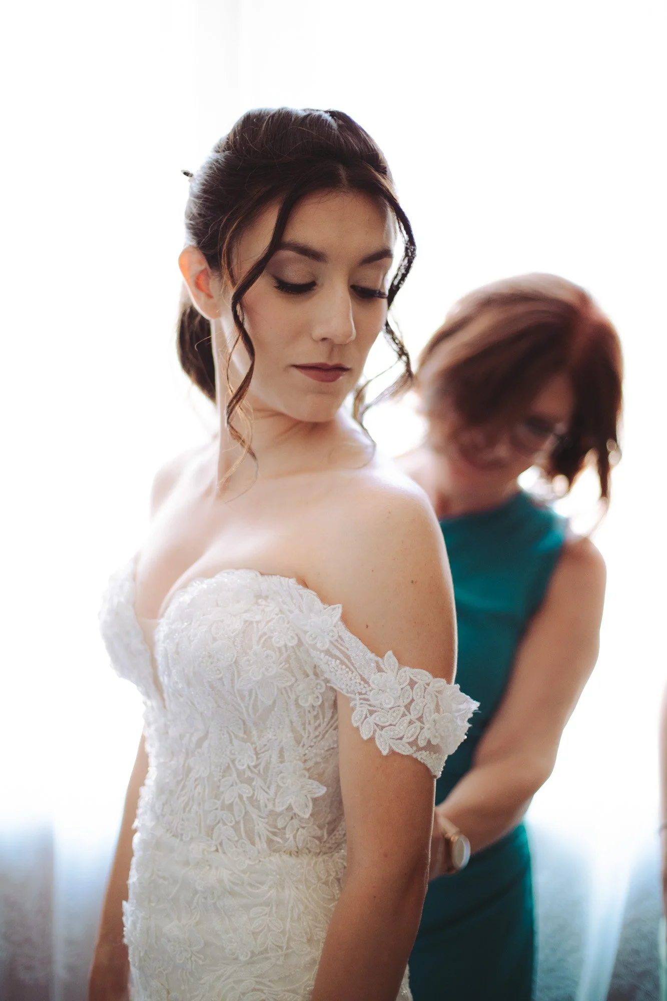 La mamma aiuta la sposa a indossare l'abito bianco in pizzo con spalle scoperte durante i preparativi