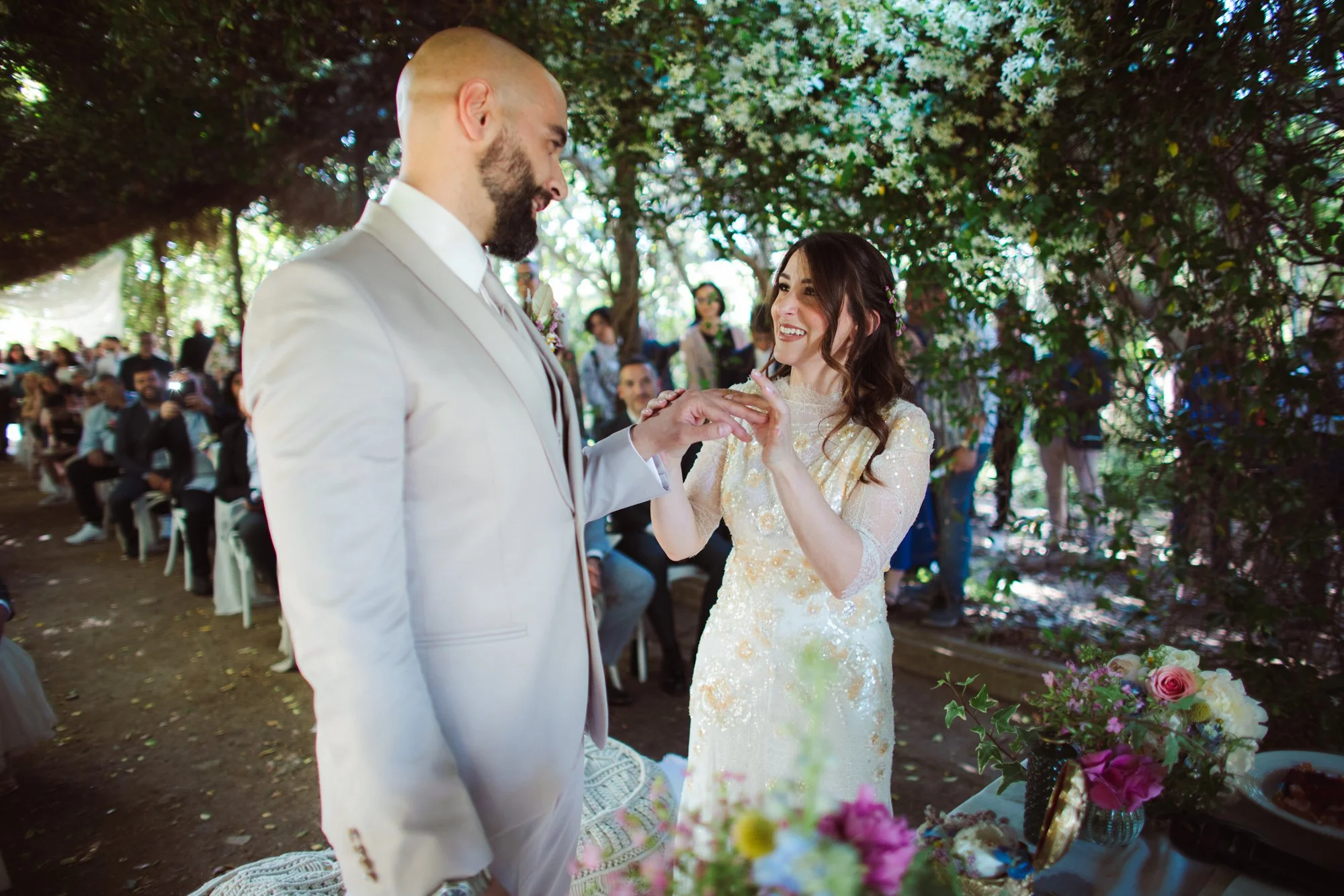 La sposa sorridente infila l'anello al dito dello sposo sotto una pergola di fiori durante la cerimonia all'aperto
