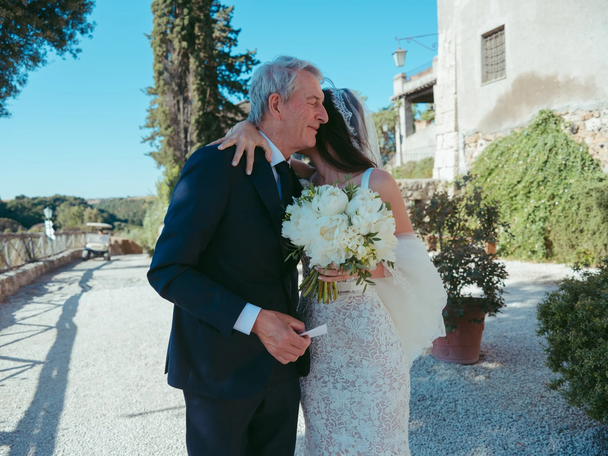 La sposa abbraccia con emozione il padre tenendo il bouquet di peonie bianche all'aperto a Borgo di Tragliata