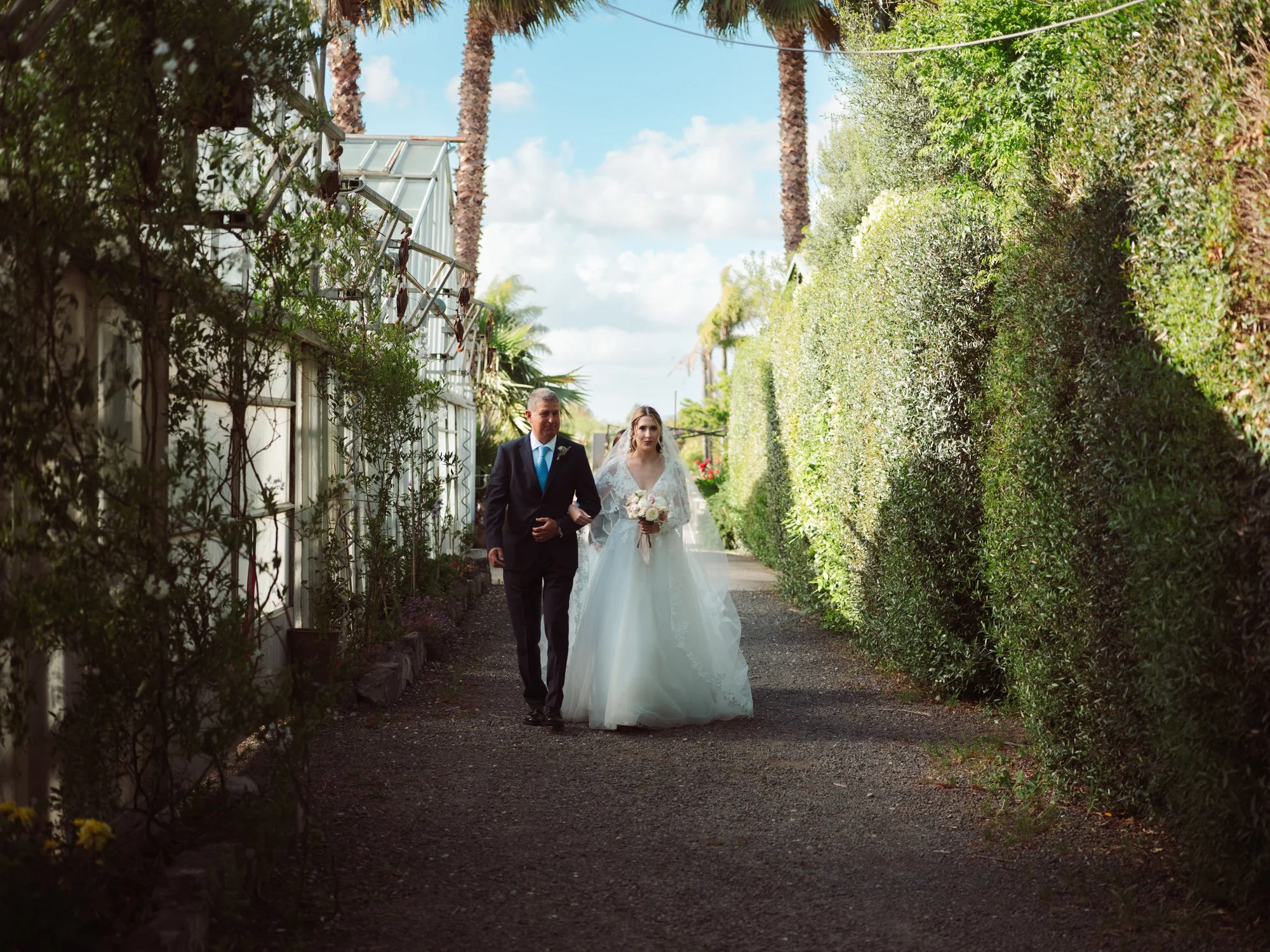 La sposa in abito bianco con velo e bouquet cammina a braccetto con il padre in abito scuro lungo il viale